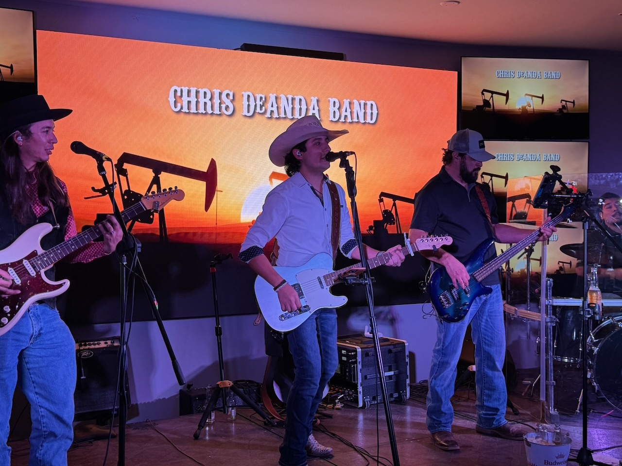 Three musicians perform on stage with guitars, wearing hats, in front of a large screen displaying 'Chris Deanda Band'.