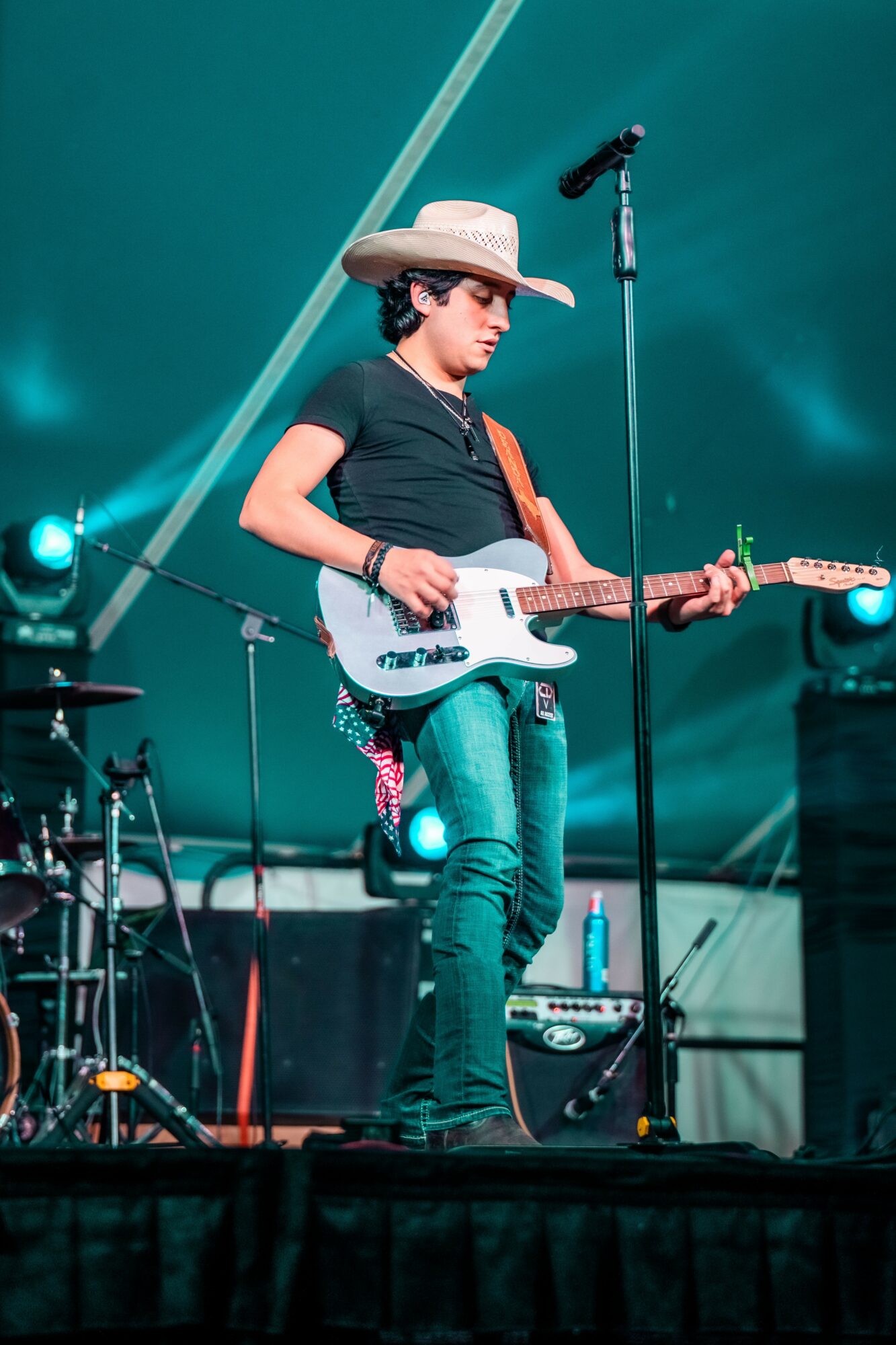 Young person playing electric guitar on stage, wearing a wide-brimmed hat and black t-shirt, with musical equipment around.