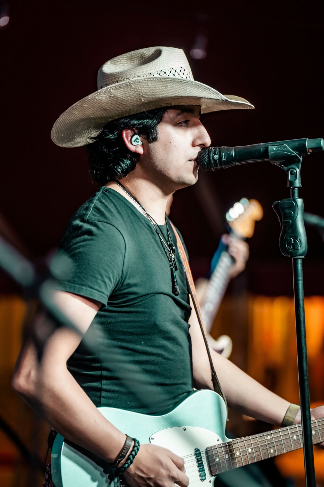 Young man wearing a cowboy hat singing into a microphone while playing an electric guitar, with a blurred background.