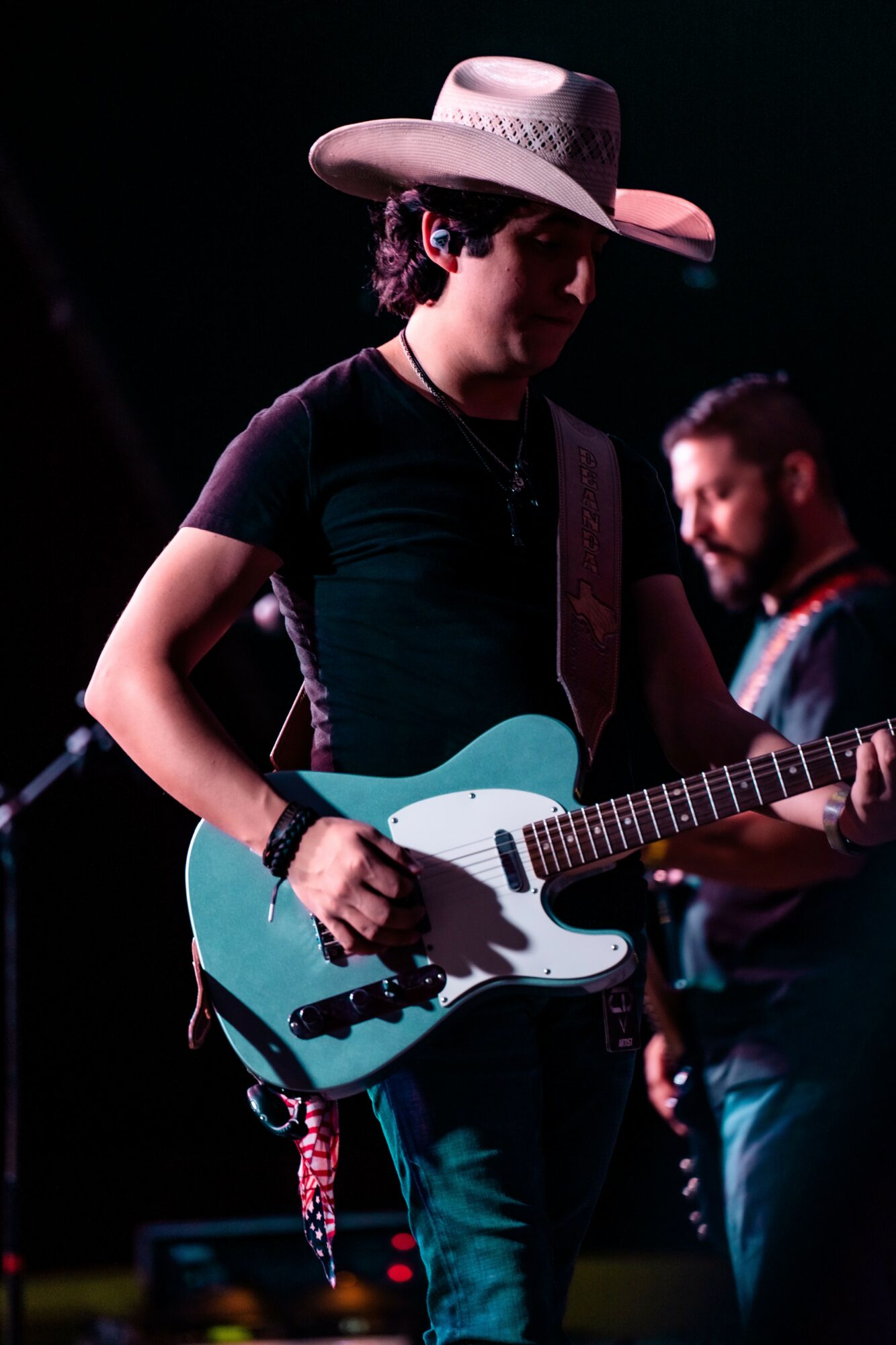 Person wearing a wide-brimmed hat playing an electric guitar on stage with another musician in the background.