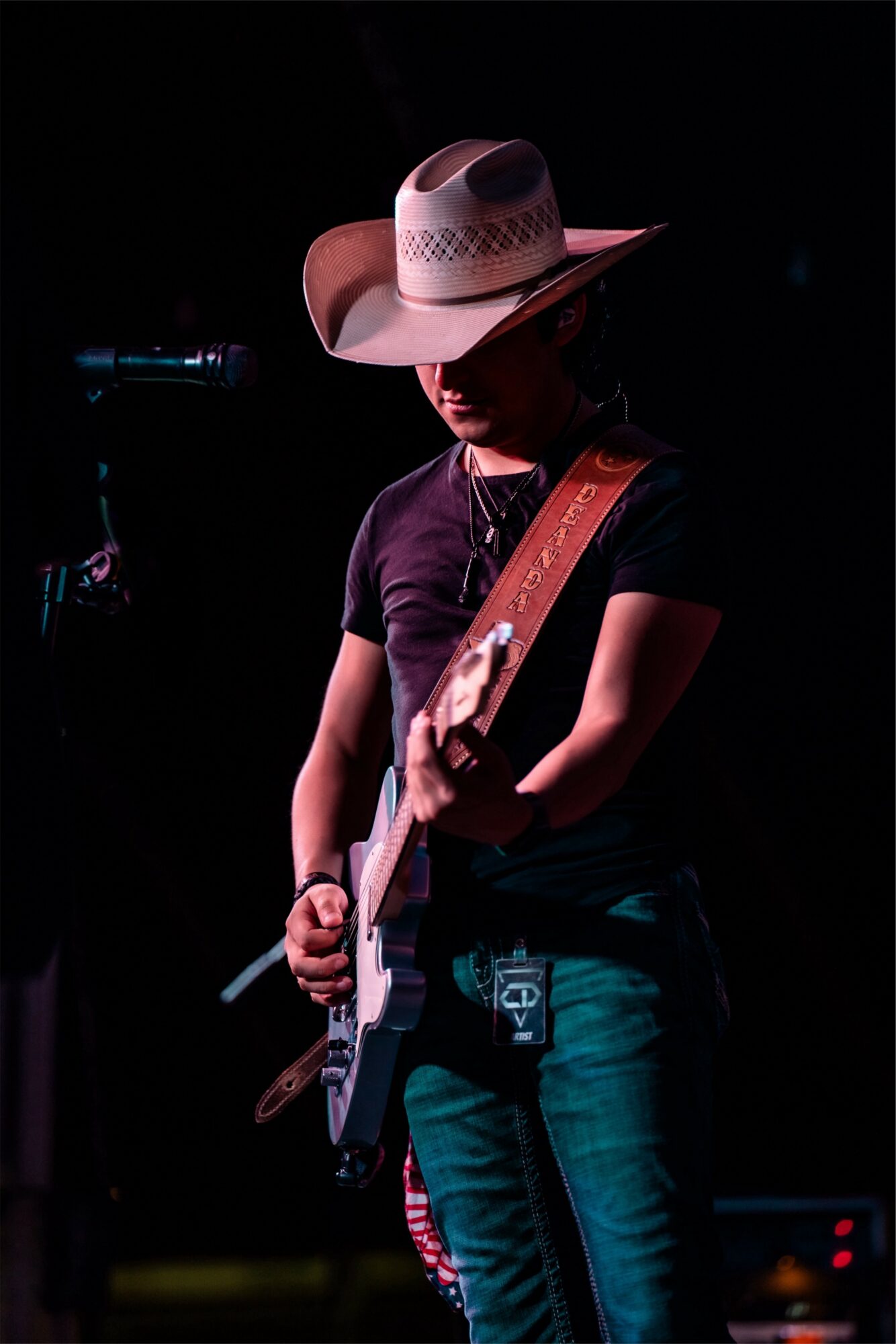 Person wearing a cowboy hat playing an electric guitar on stage, dark background, focused expression.