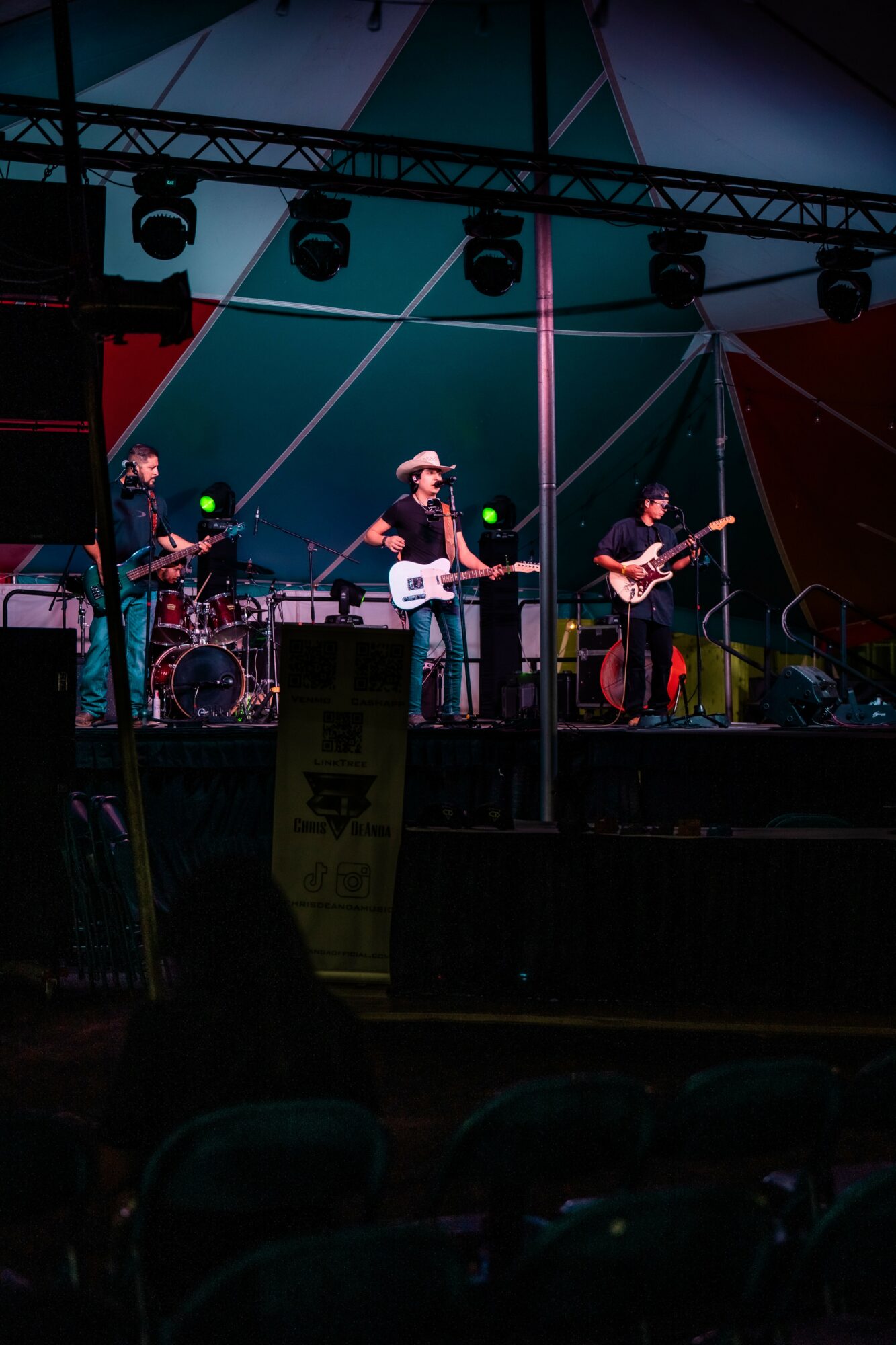 Band performing on stage with four musicians, colorful geometric backdrop, stage lights, and musical instruments.