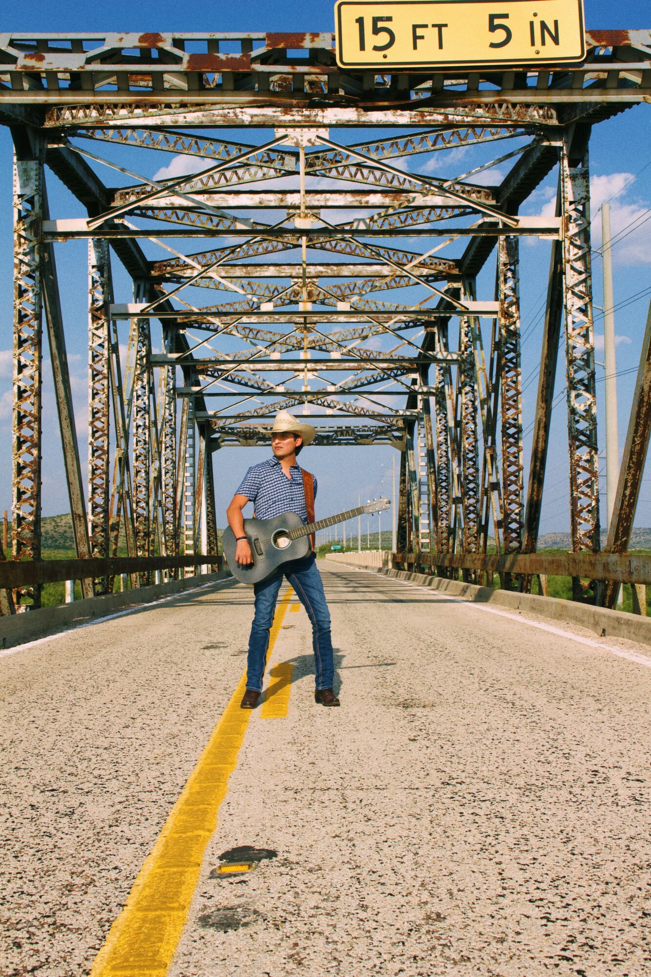 Person standing in the middle of a bridge with a guitar, metal structure overhead, and a height restriction sign above, clear sky