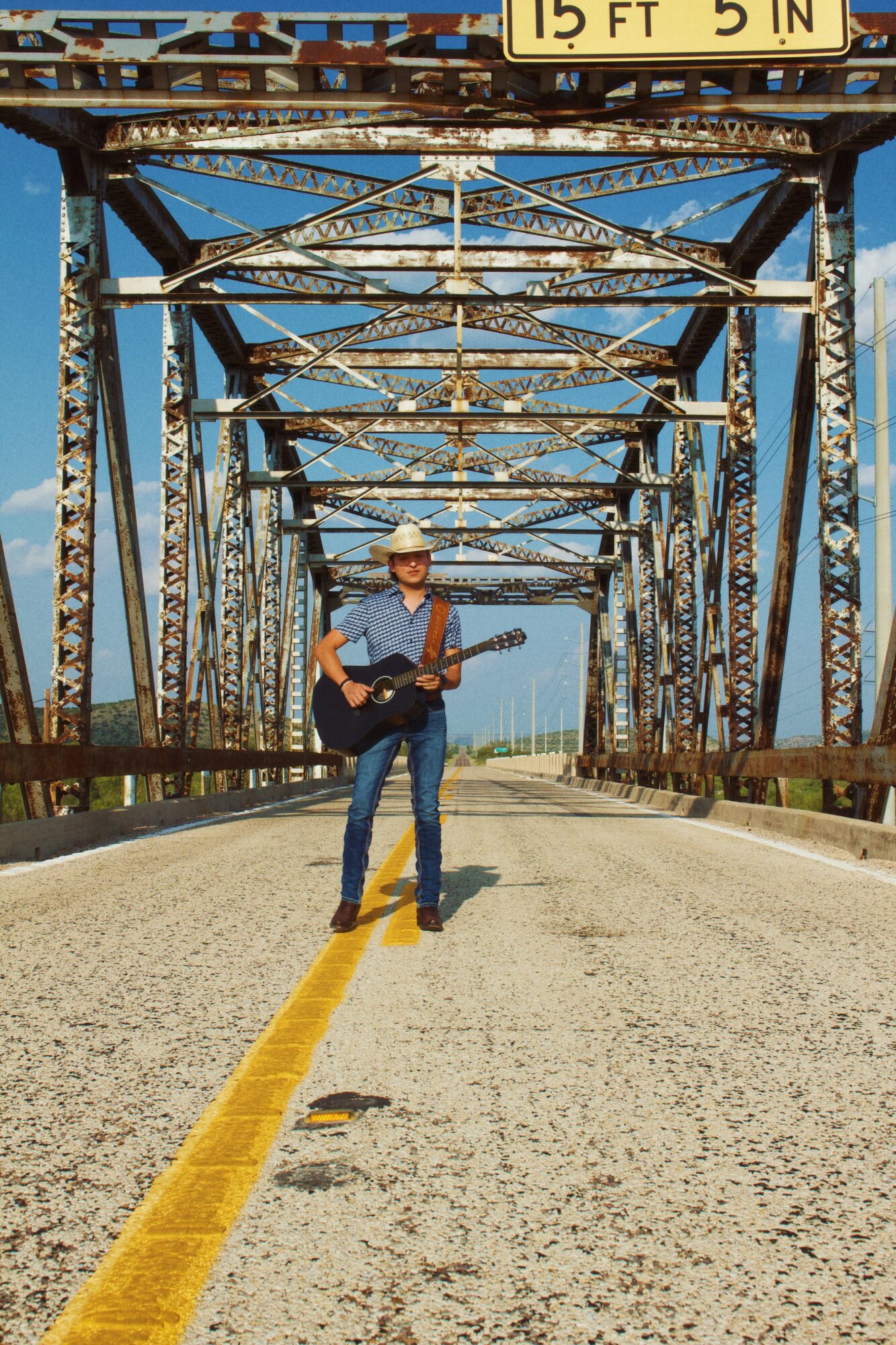 Person wearing a hat playing guitar standing on a road under a metal bridge structure.