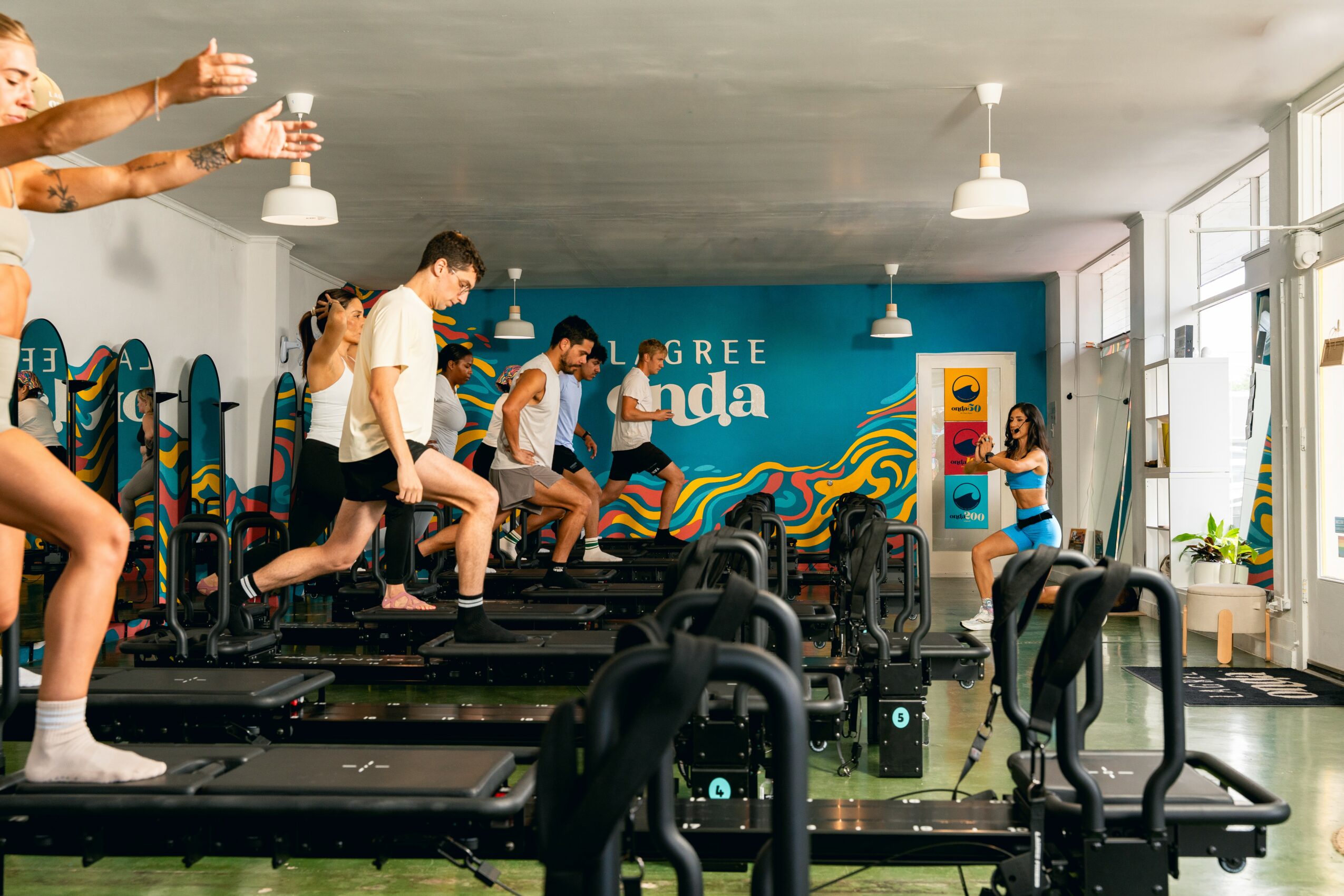 People exercising on mini trampolines in a fitness studio with a blue wall and colorful mural, sunlight coming through windows.