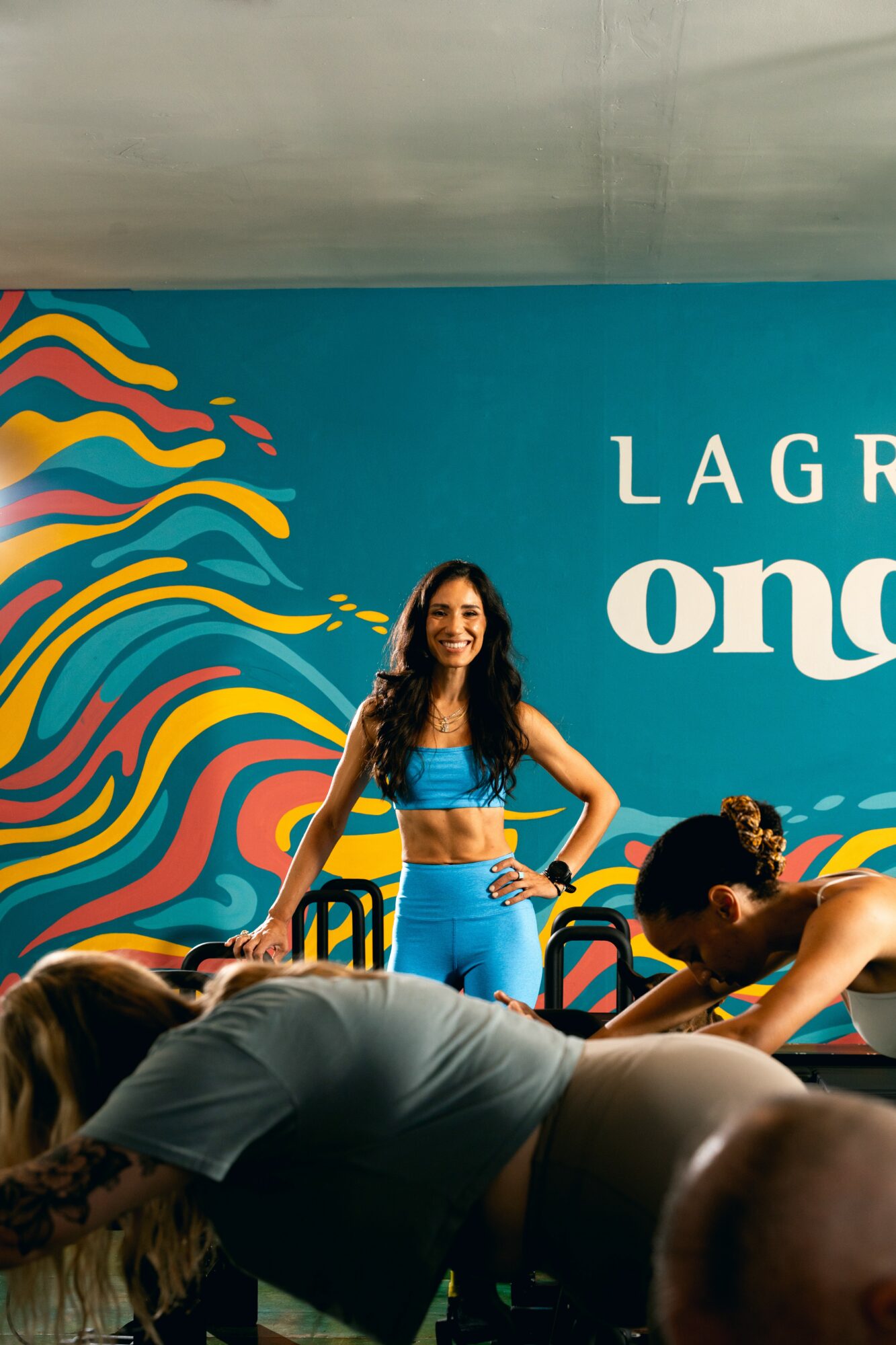 Woman standing with hands on hips in front of colorful mural, smiling, with people stretching in foreground.