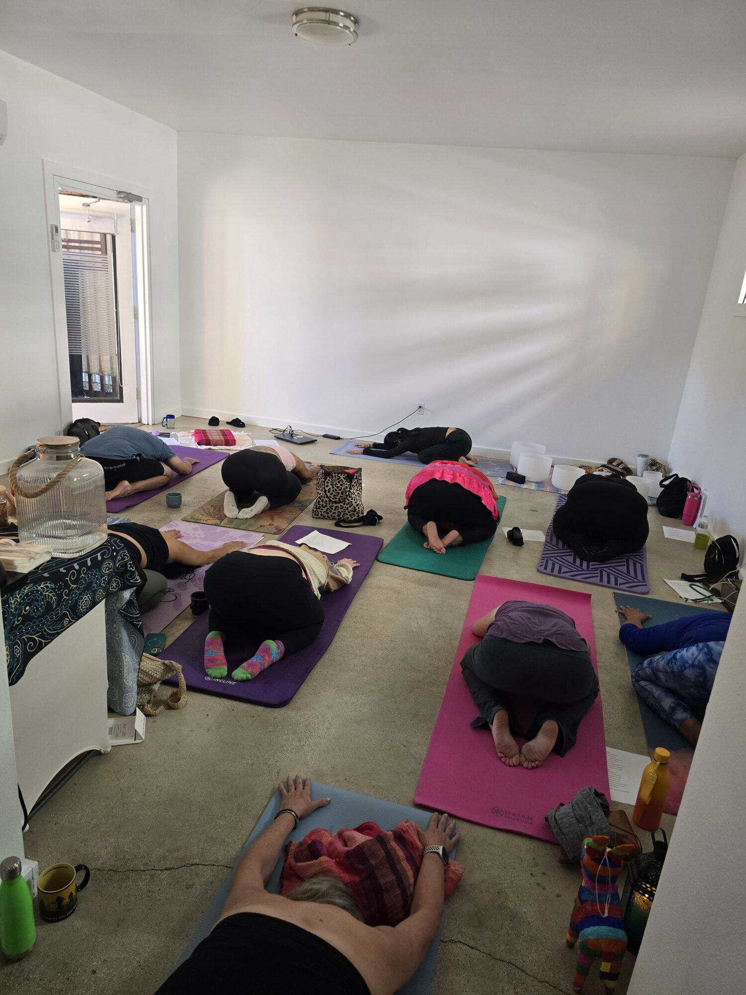 Group of people practicing yoga on mats in a bright room with white walls and a window, some with hands in prayer position.