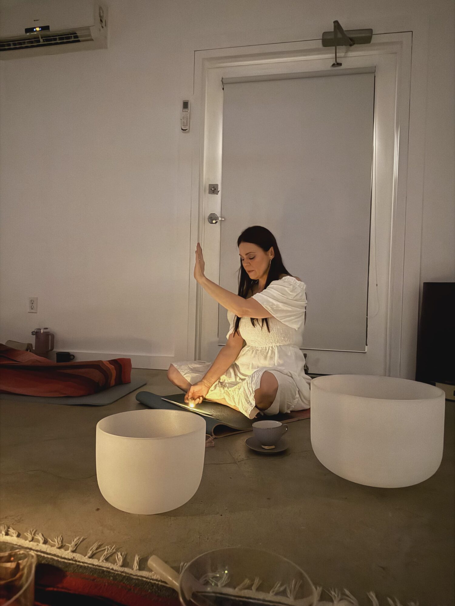 Woman sitting on the floor reading a book, surrounded by large white bowls, in a minimalistic room with a door behind her.