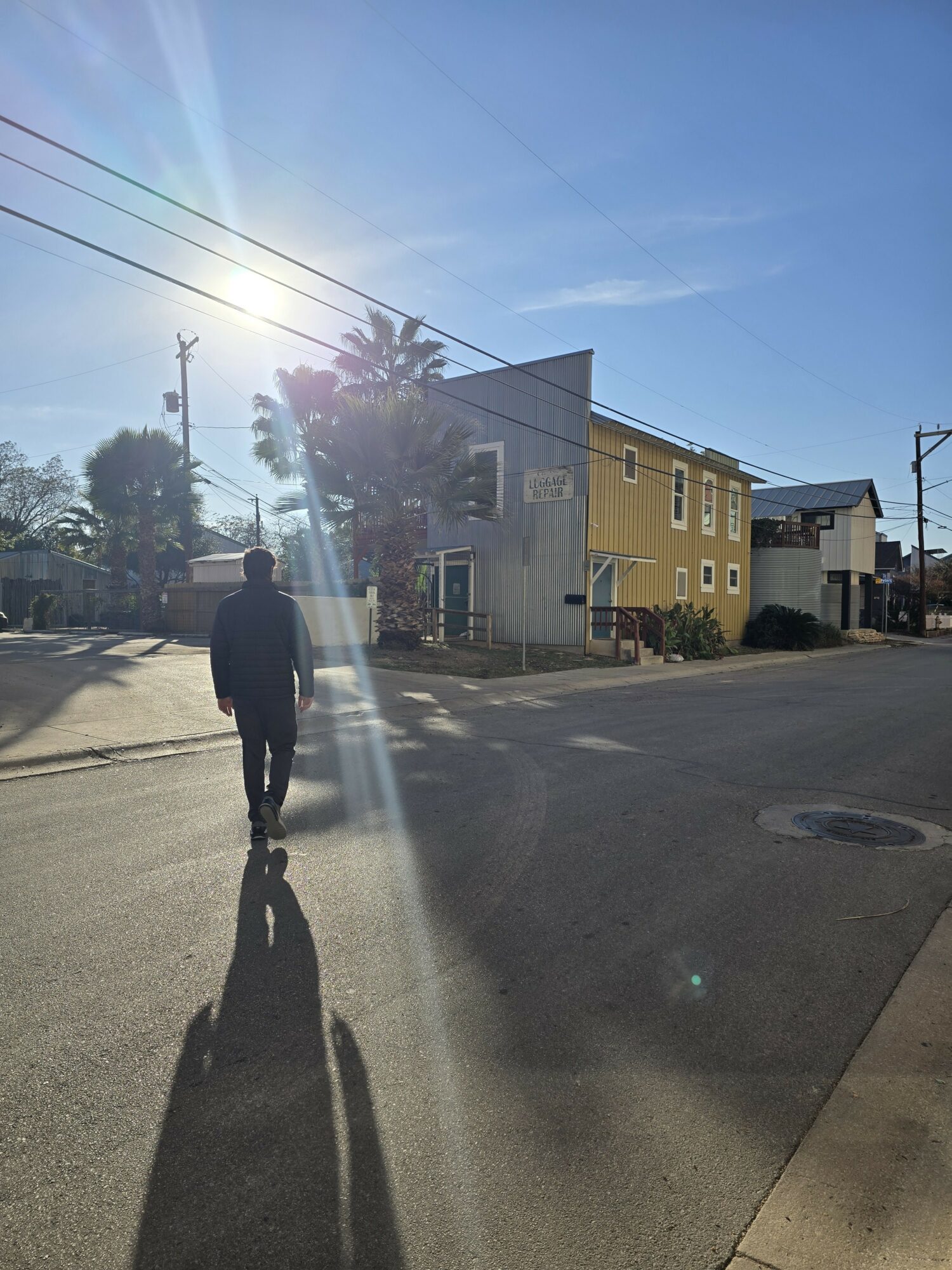 Person walking on street with sunlight shining, buildings and trees in background, clear blue sky.