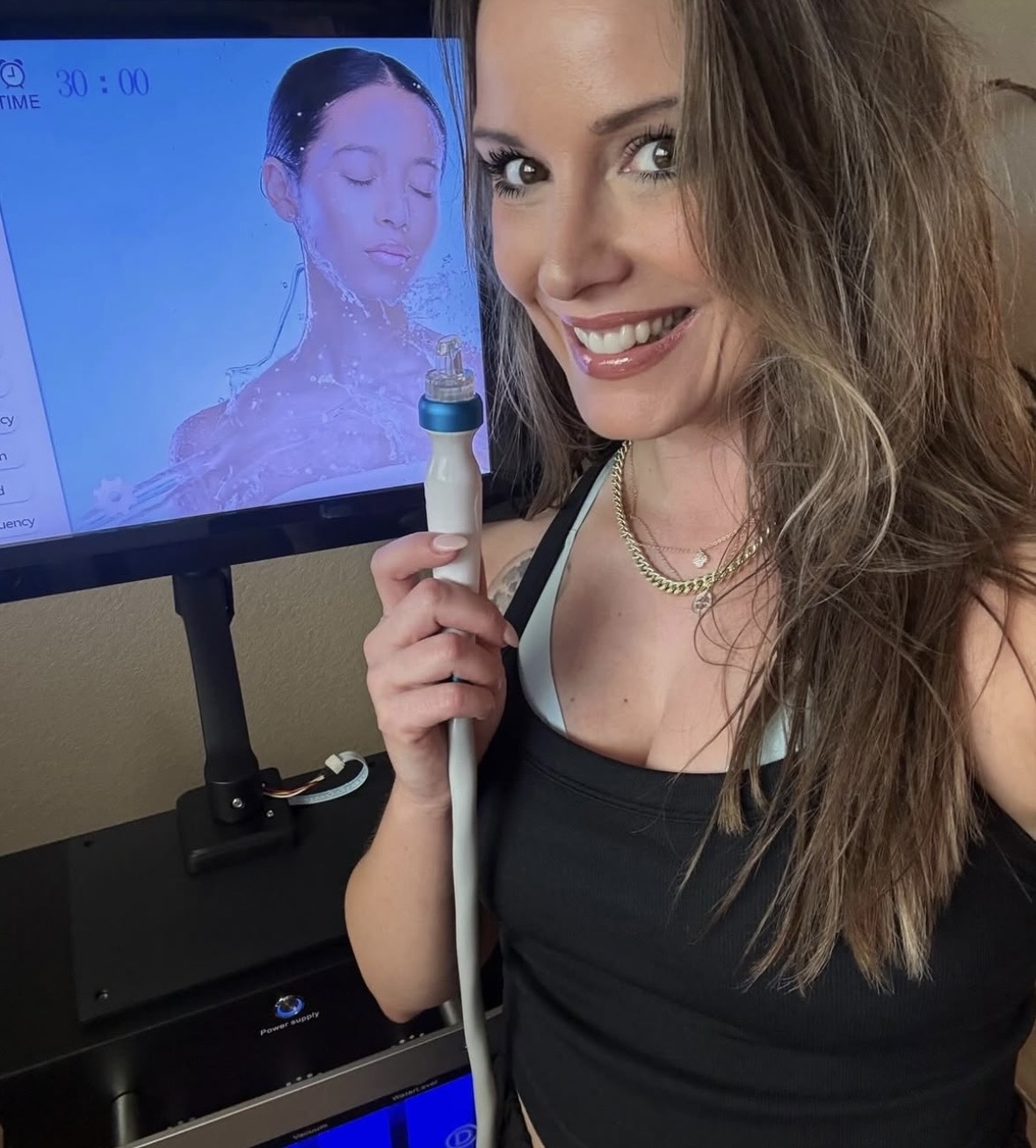 Woman smiling holding a skincare device in front of a monitor displaying her face with closed eyes.