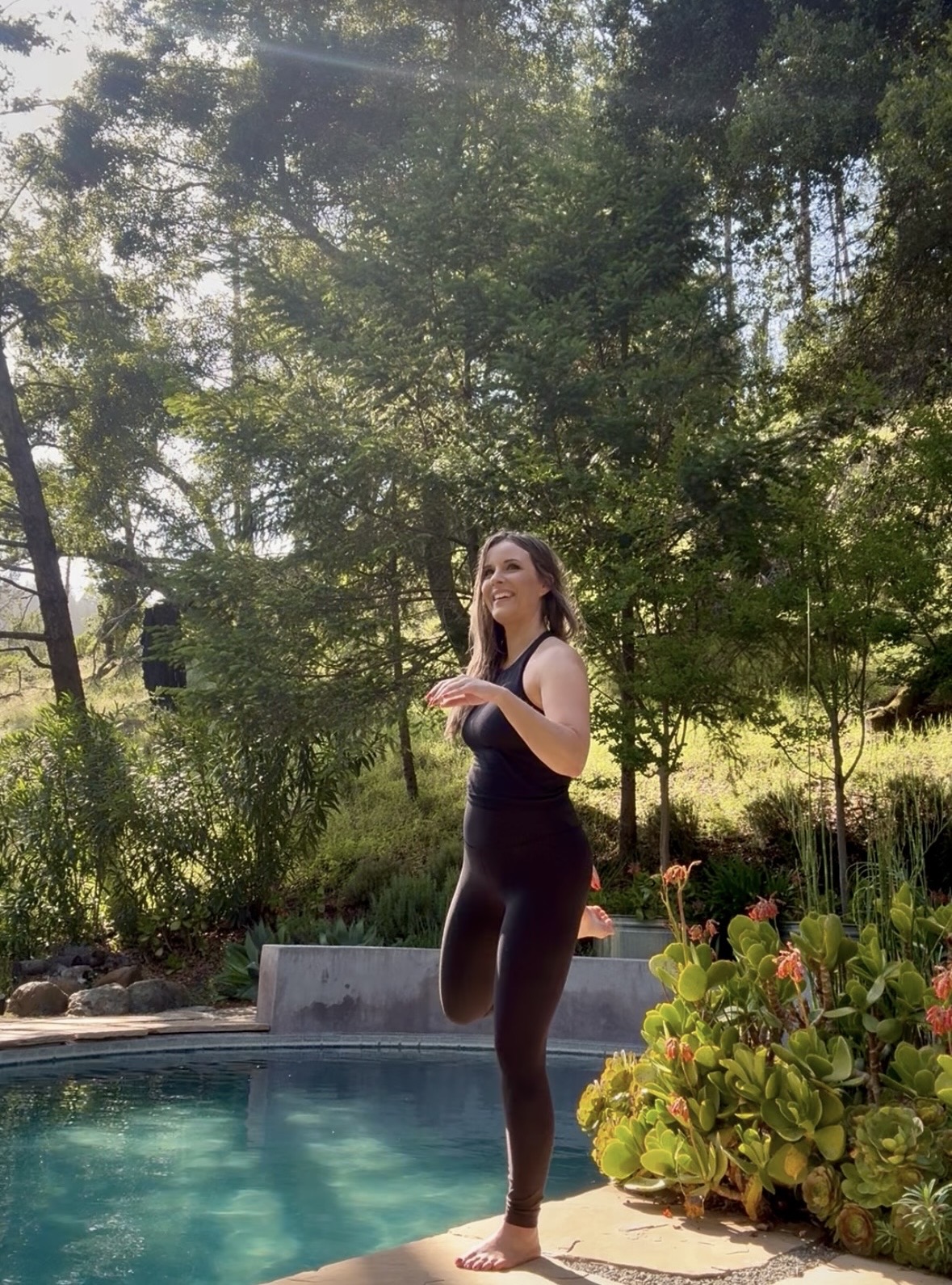 Woman standing by a pool surrounded by trees and plants outdoors, smiling and looking to the side.