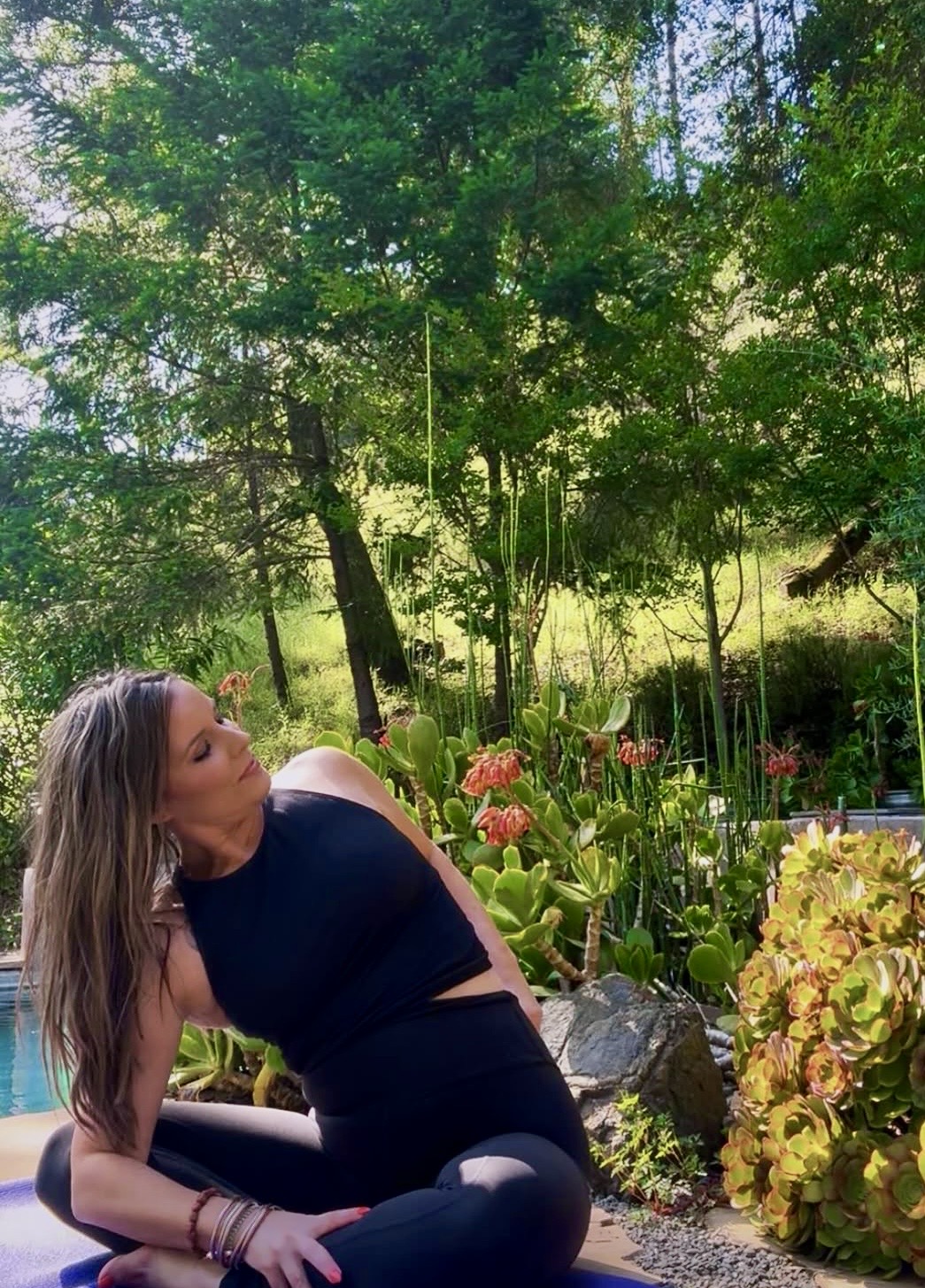 Woman sitting outdoors among trees and plants, wearing a black top and leggings, looking to the side.