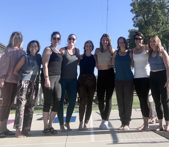 Group of nine women standing outdoors on a sunny day, smiling, with trees and a building in the background.