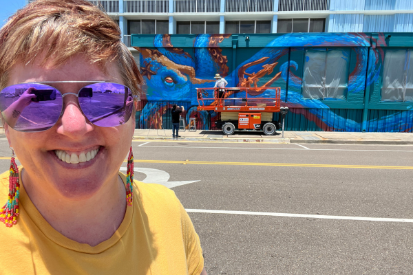 Smiling person with purple sunglasses and colorful earrings in front of a mural with an octopus, on a city street.