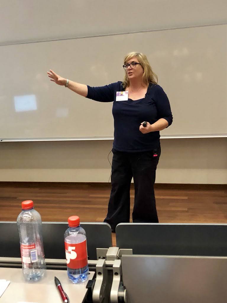 Woman standing in front of a whiteboard, gesturing with her right arm, wearing glasses and dark clothing, in a classroom or conference room.