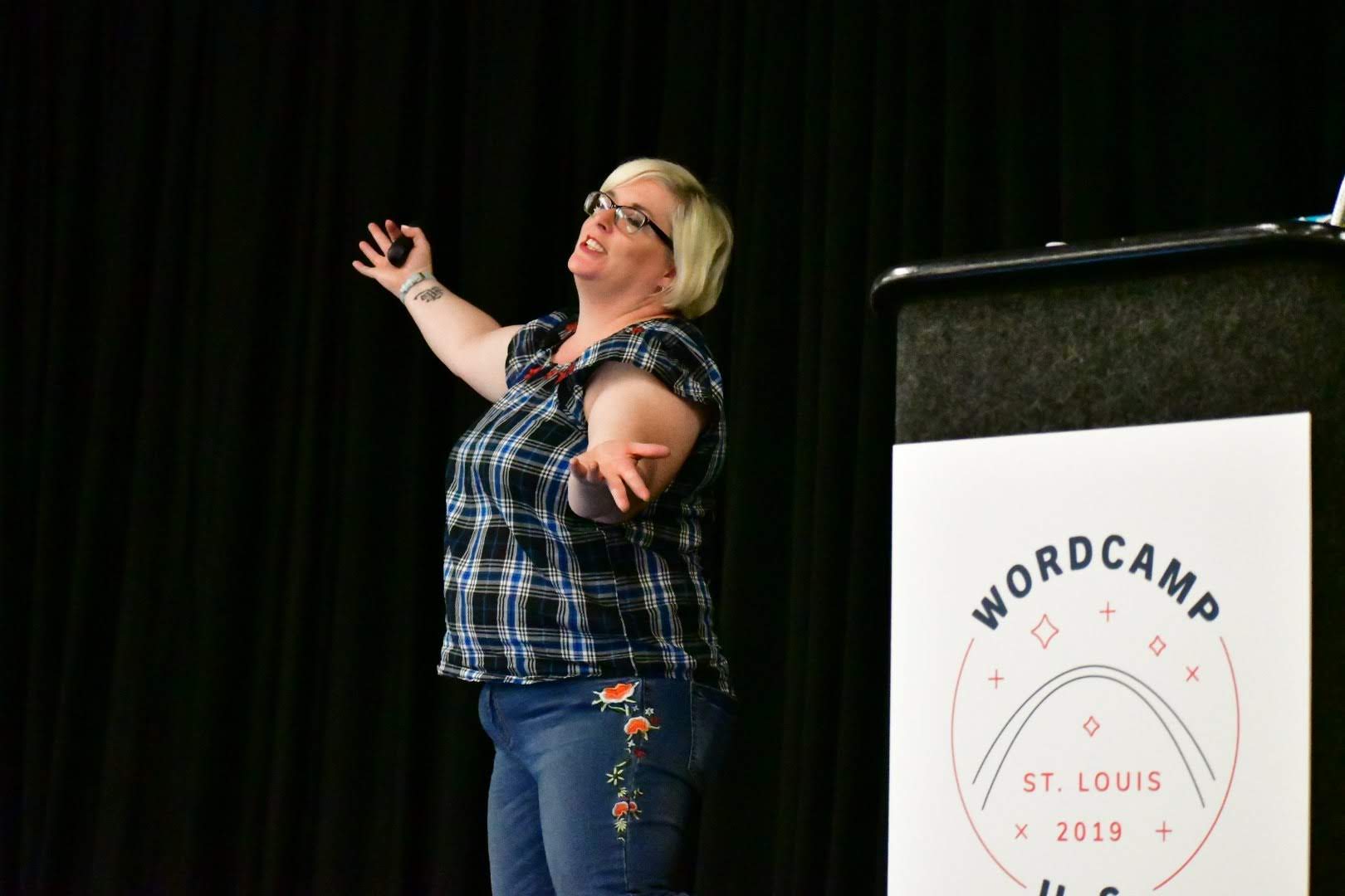 Woman speaking on stage with black curtain background, gesturing with hands, standing near a podium with WordCamp sign.