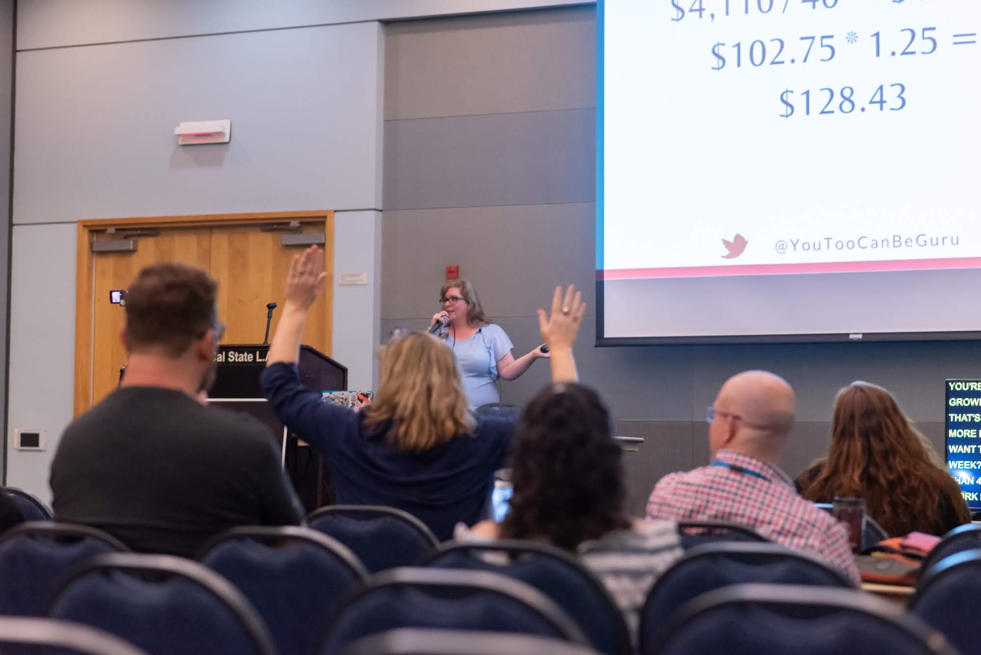 Person with blonde hair raising hand in a conference room with audience and presentation screen.