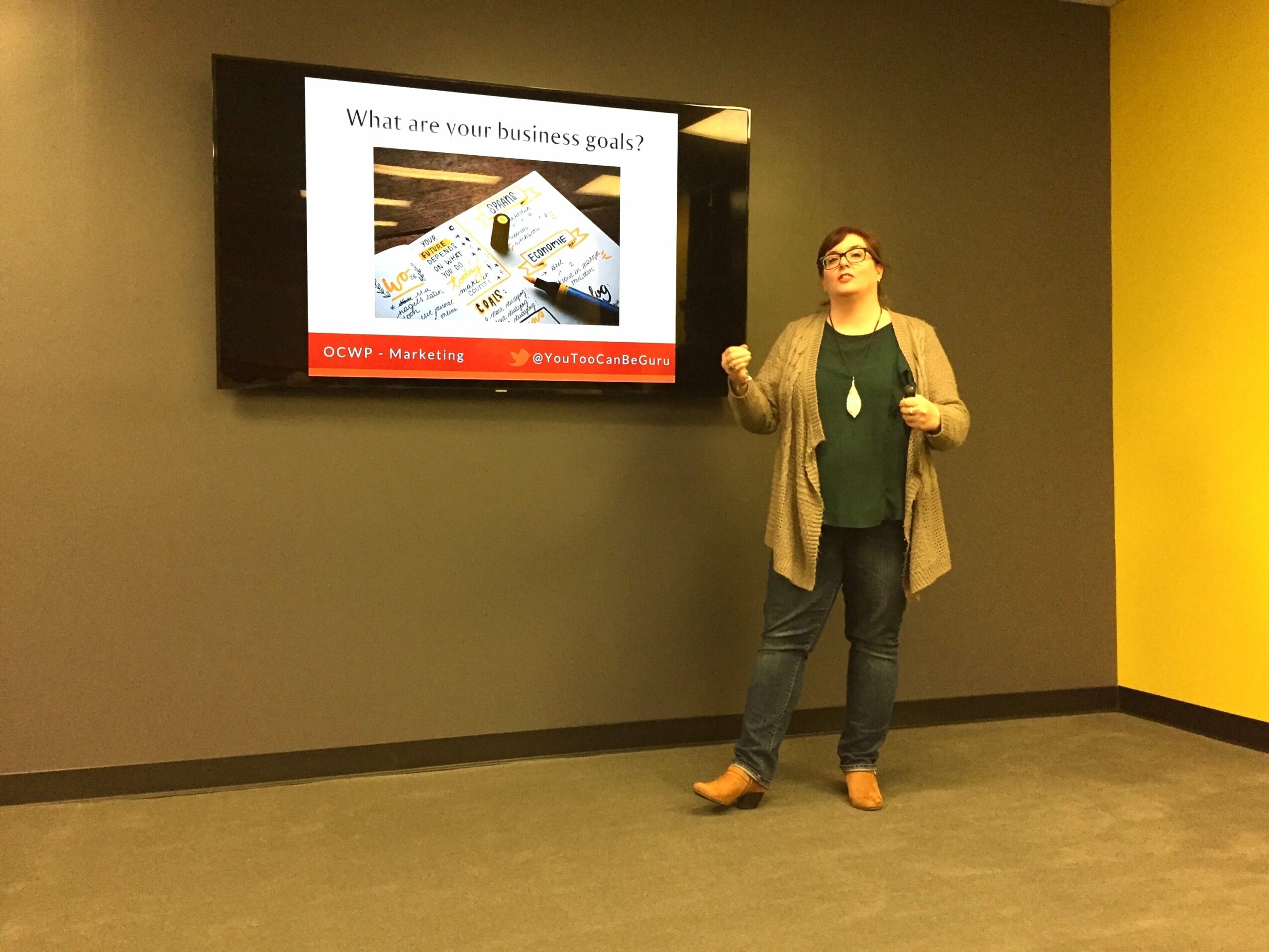 Woman standing in front of a screen displaying a presentation slide about business goals, in a room with green and yellow walls.
