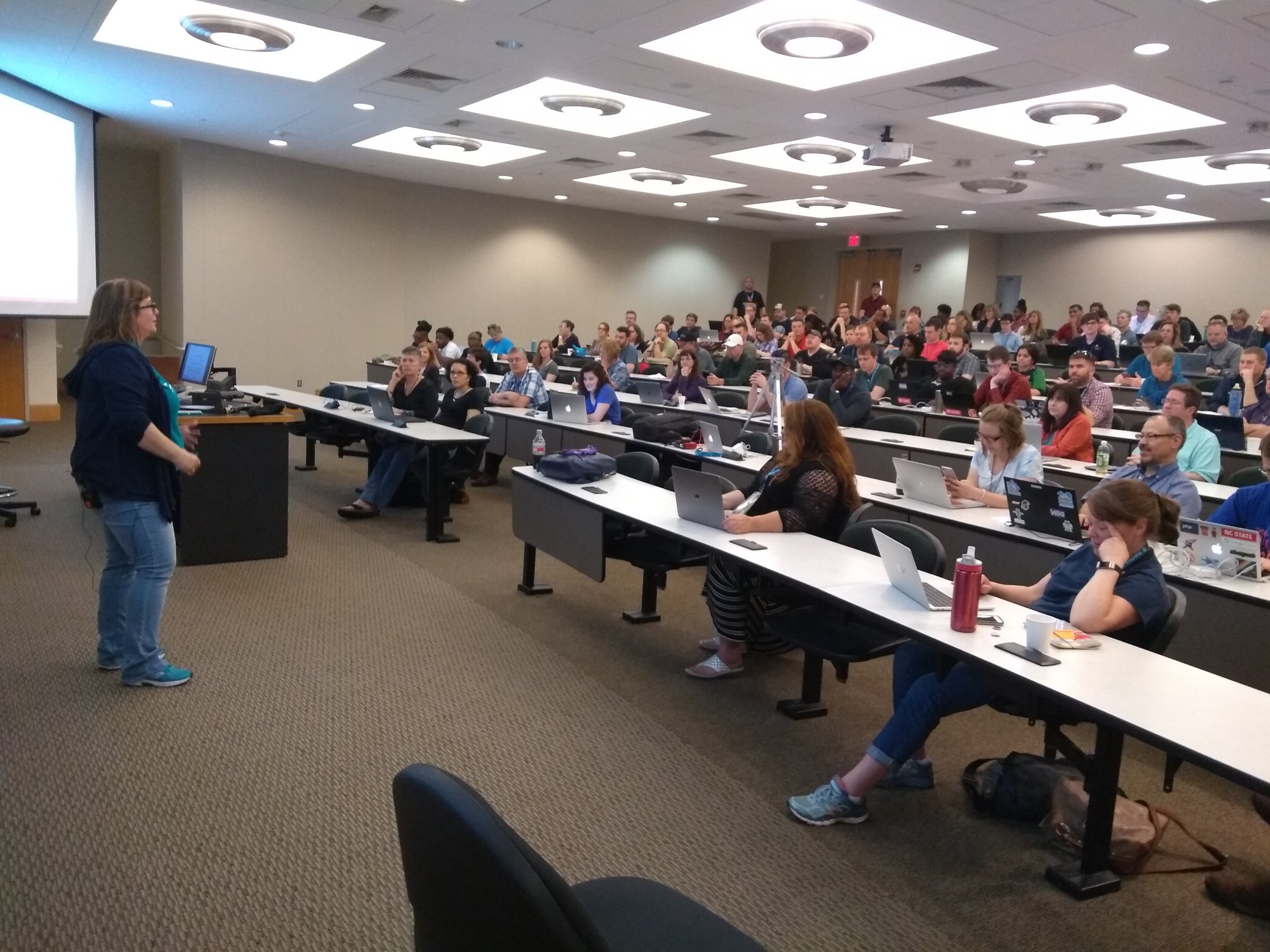 A woman stands at the front of a classroom with students seated at desks, some using laptops, in a large room with ceiling lights.