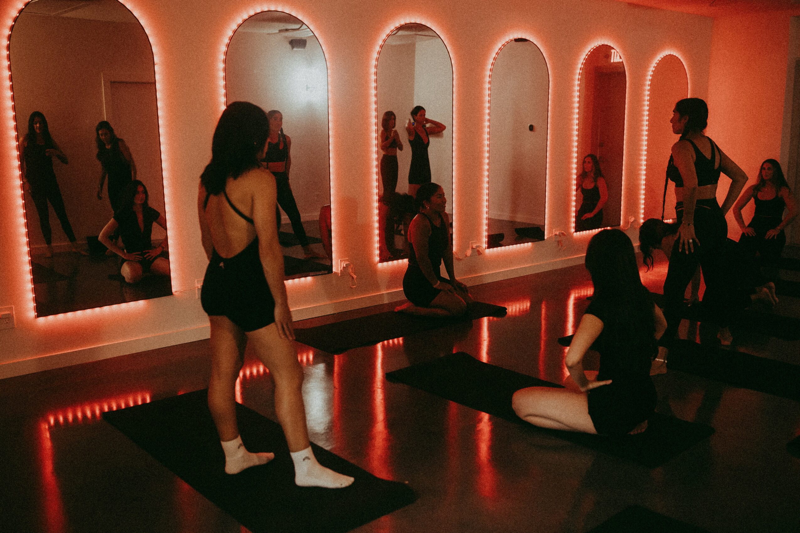 Group of people in a dance or yoga class in a room with illuminated mirrors, some sitting and some standing, with black mats.