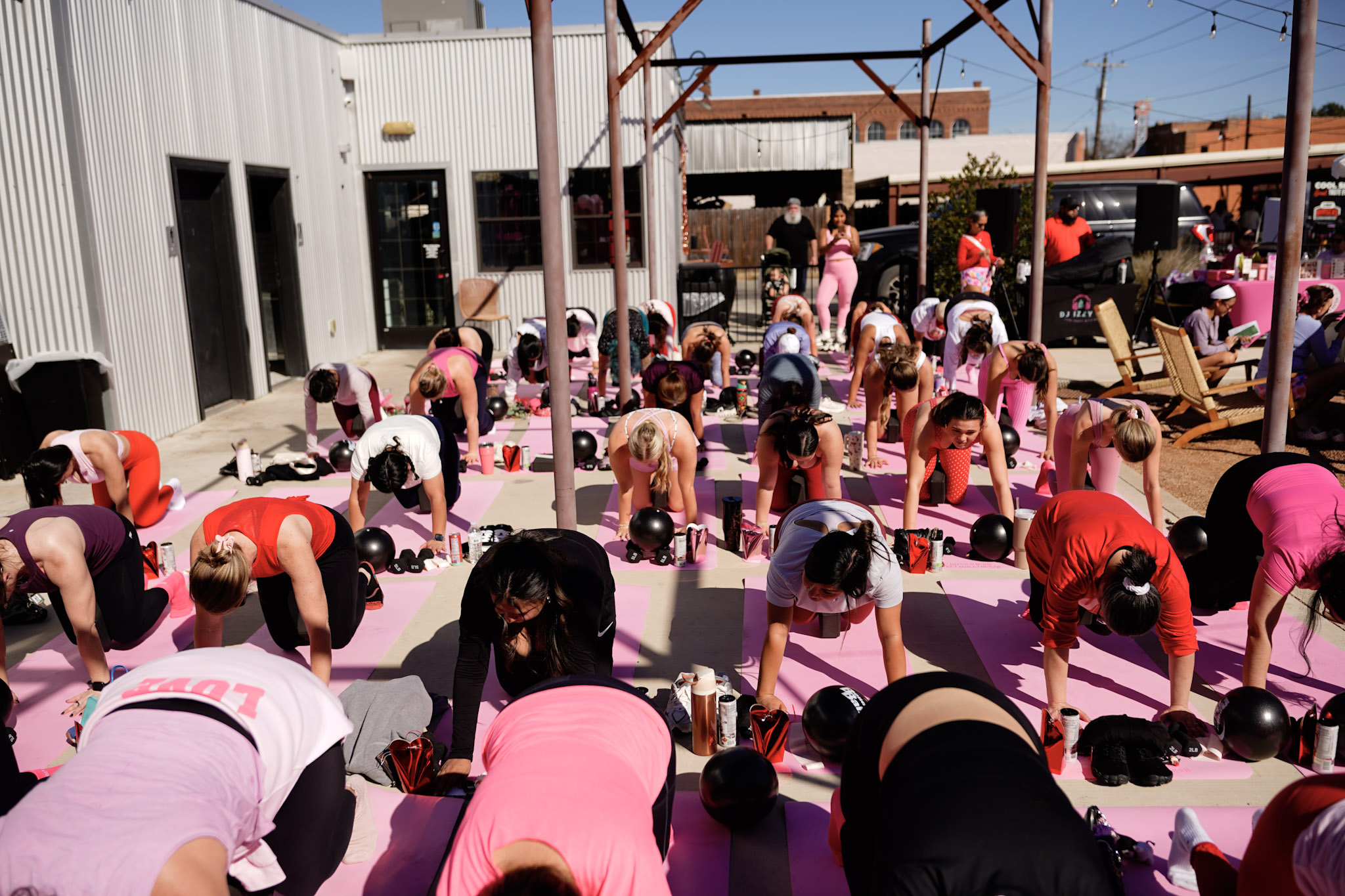 Group of people participating in outdoor fitness class, bending forward on pink mats, with gym equipment and building in background.