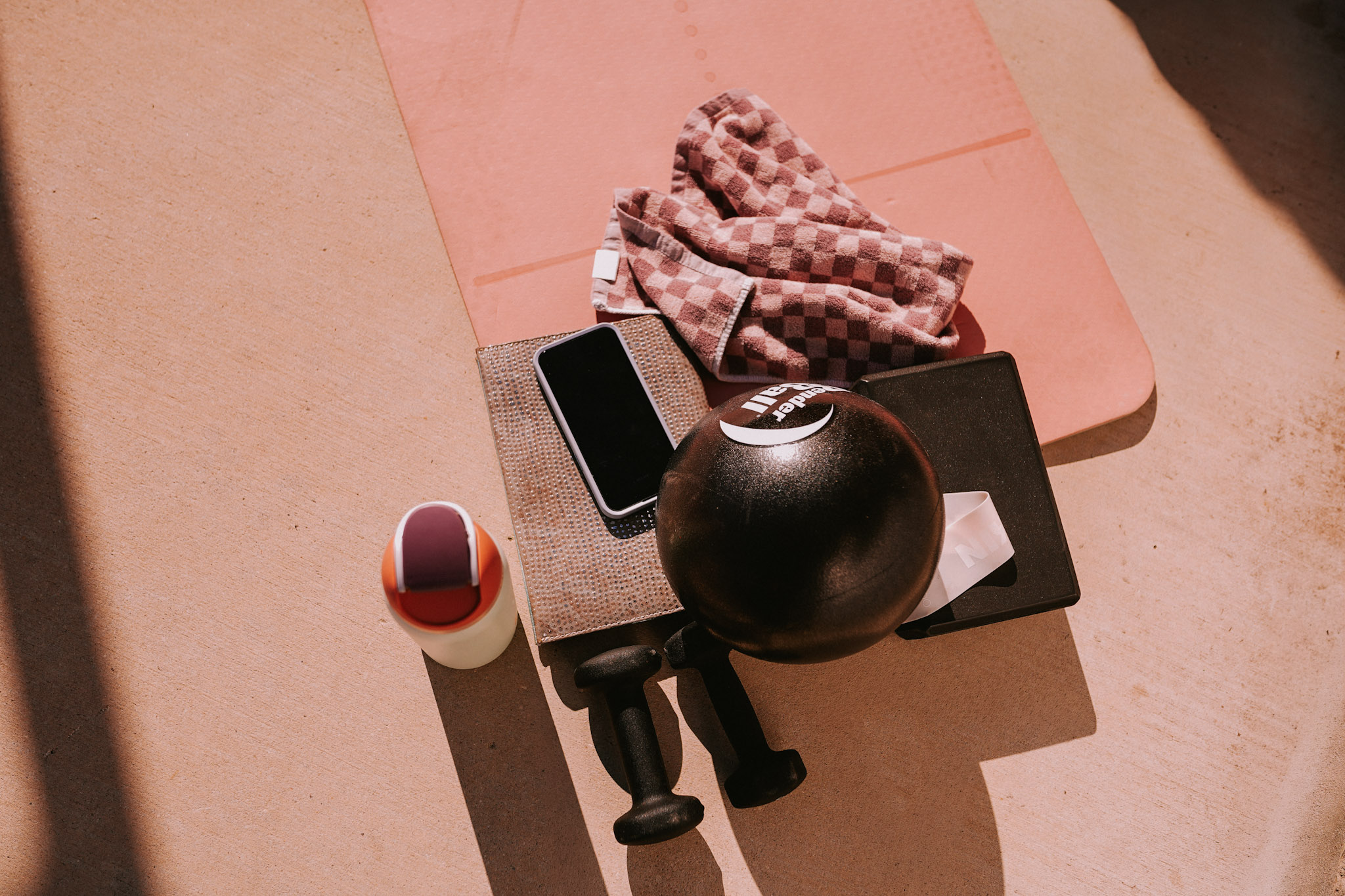 Pink table with a black ball, a small black hammer, a smartphone, a cloth, and a white cup on a pink surface.