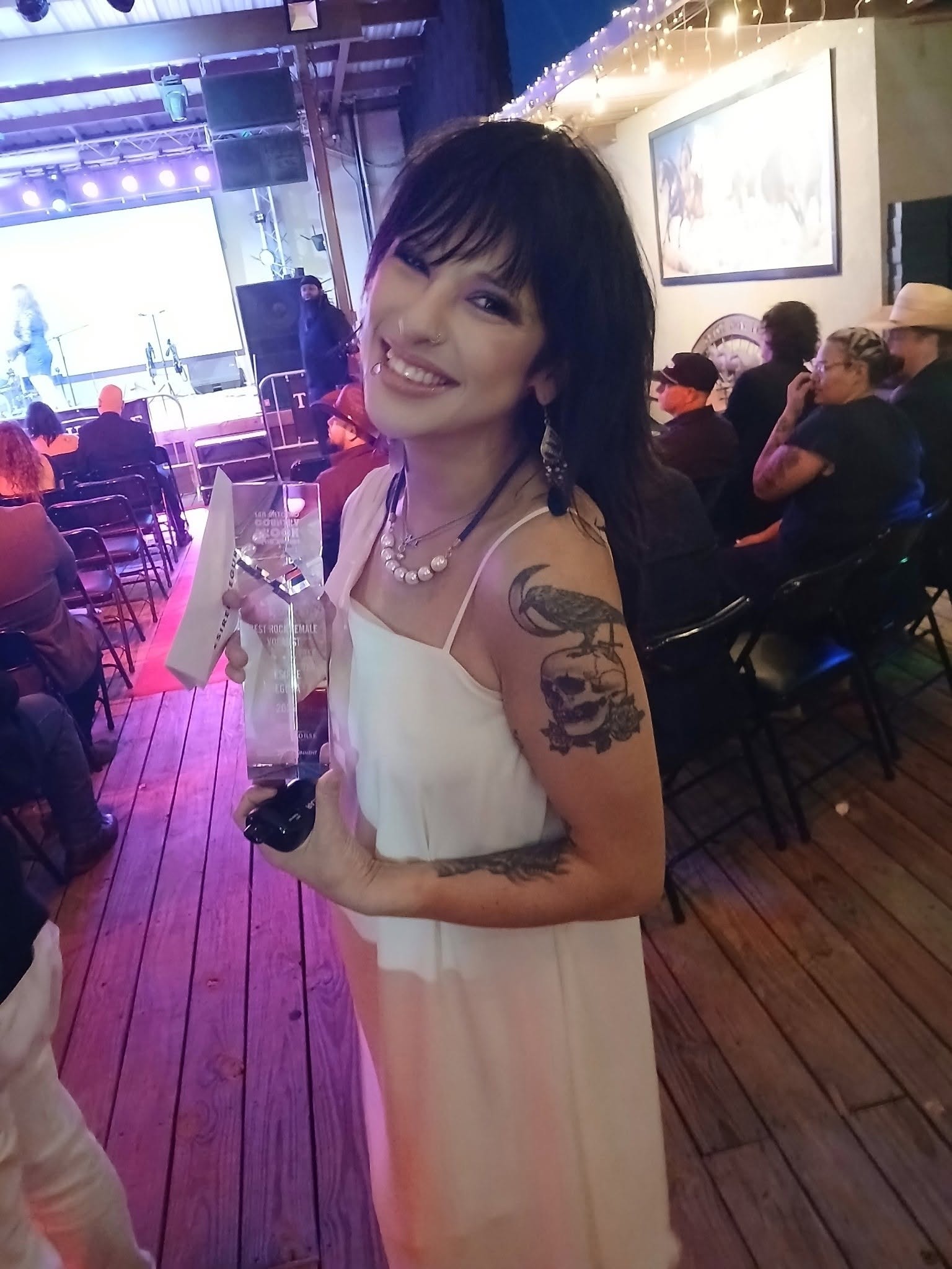 Woman with short dark hair in a white dress holding a drink, smiling at an indoor event with seated audience.