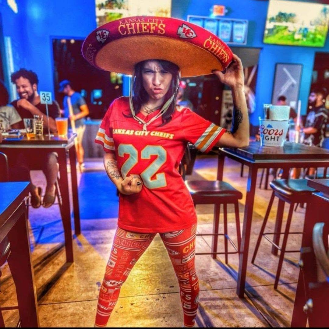 Person wearing a Kansas City Chiefs jersey and leggings, holding a large Chiefs hat in a lively bar setting.