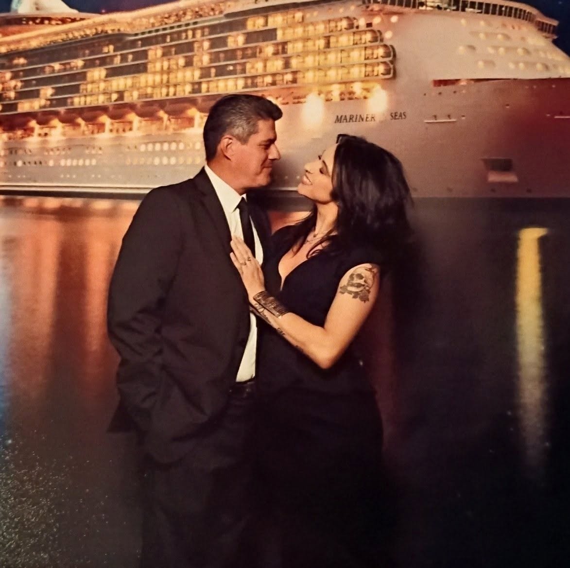 A man and woman face each other near a large cruise ship, with water reflecting the scene behind them.