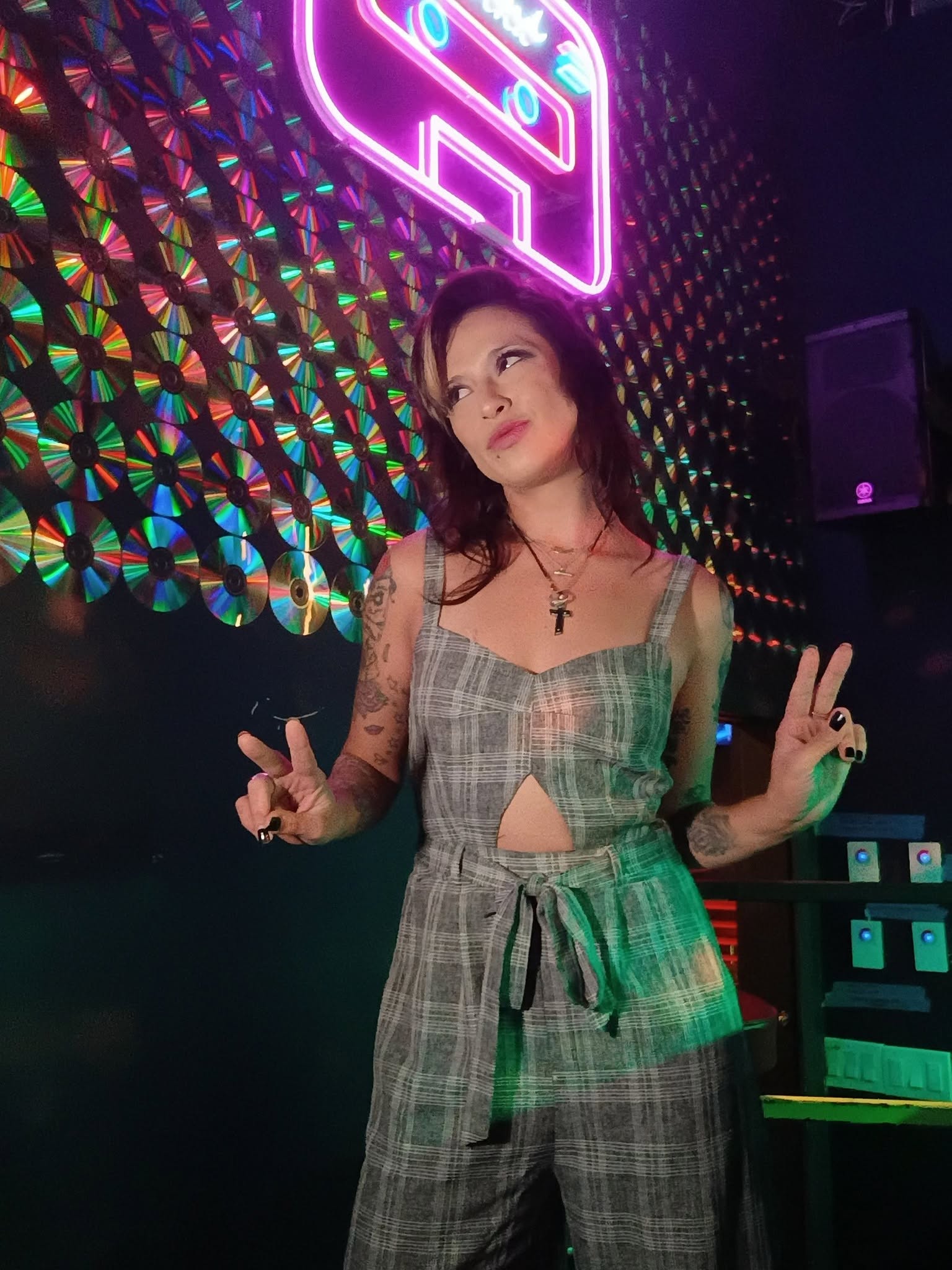 Young woman with dark hair in a plaid jumpsuit making peace signs, neon sign and colorful wall behind her.