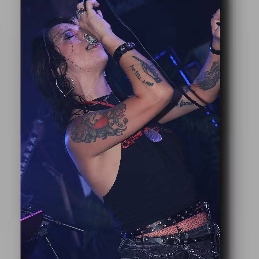 Person singing into a microphone with tattoos on arms, wearing a black sleeveless top, in a dark setting.
