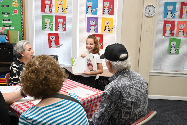 Group of people sitting at a table with a young girl standing and speaking, colorful artwork on the wall, clock on the wall.
