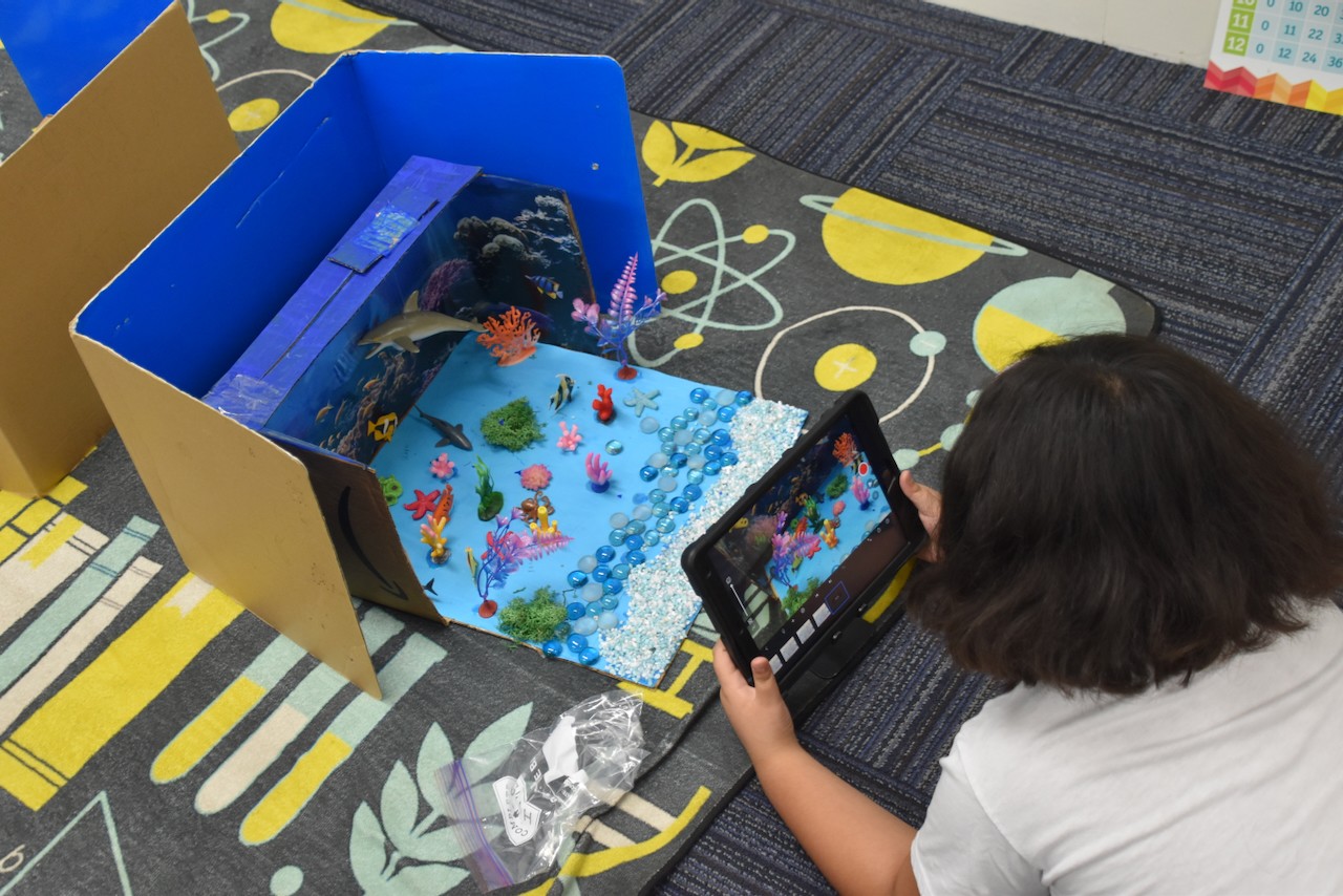 Child with dark hair and white shirt takes a photo of a colorful fish tank on a table.