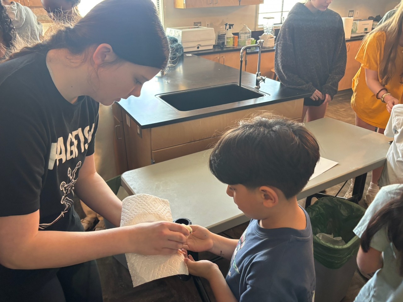 A young woman and a boy are holding hands in a classroom with a sink and other students in the background.
