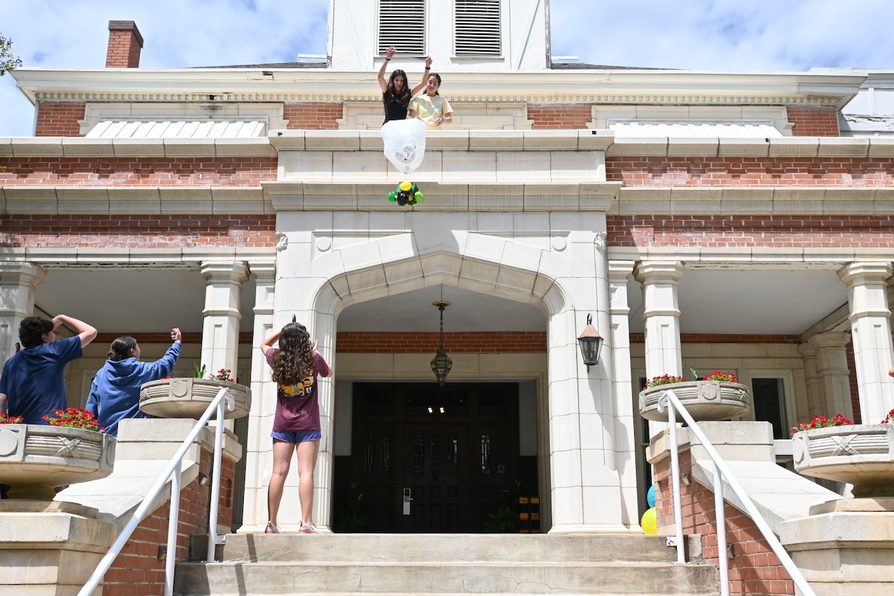 Person standing on a ledge above a building entrance, with two children on stairs below pointing upward, in front of a large building.