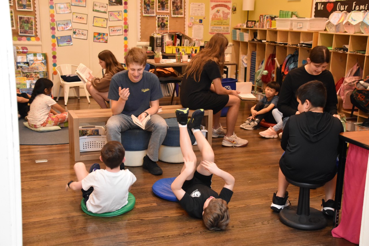 Children and adults sit on the floor and chairs in a classroom, engaging in activities and conversations.