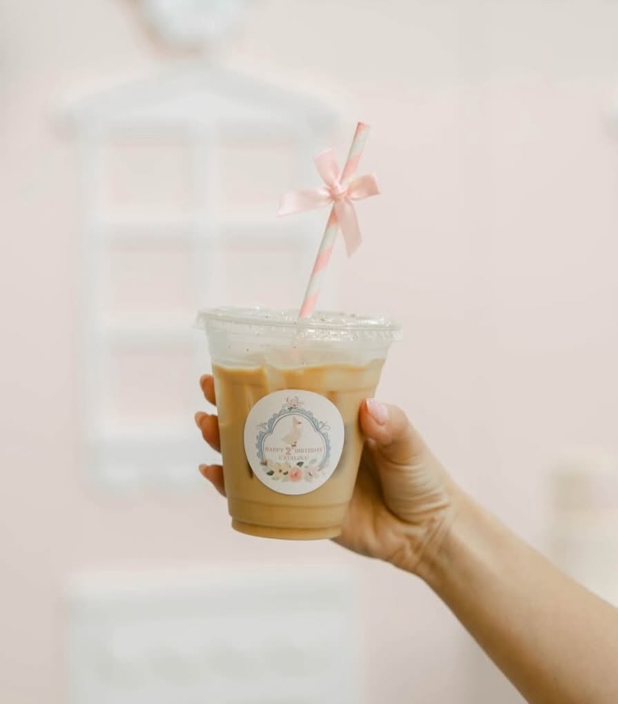 Hand holding a plastic cup with iced coffee, pink ribbon straw, and a round sticker, against a blurred background.