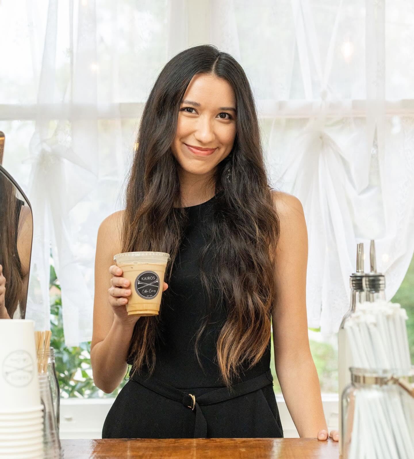 Young woman with long dark hair smiling and holding a cup of coffee, standing indoors near a window with white curtains.