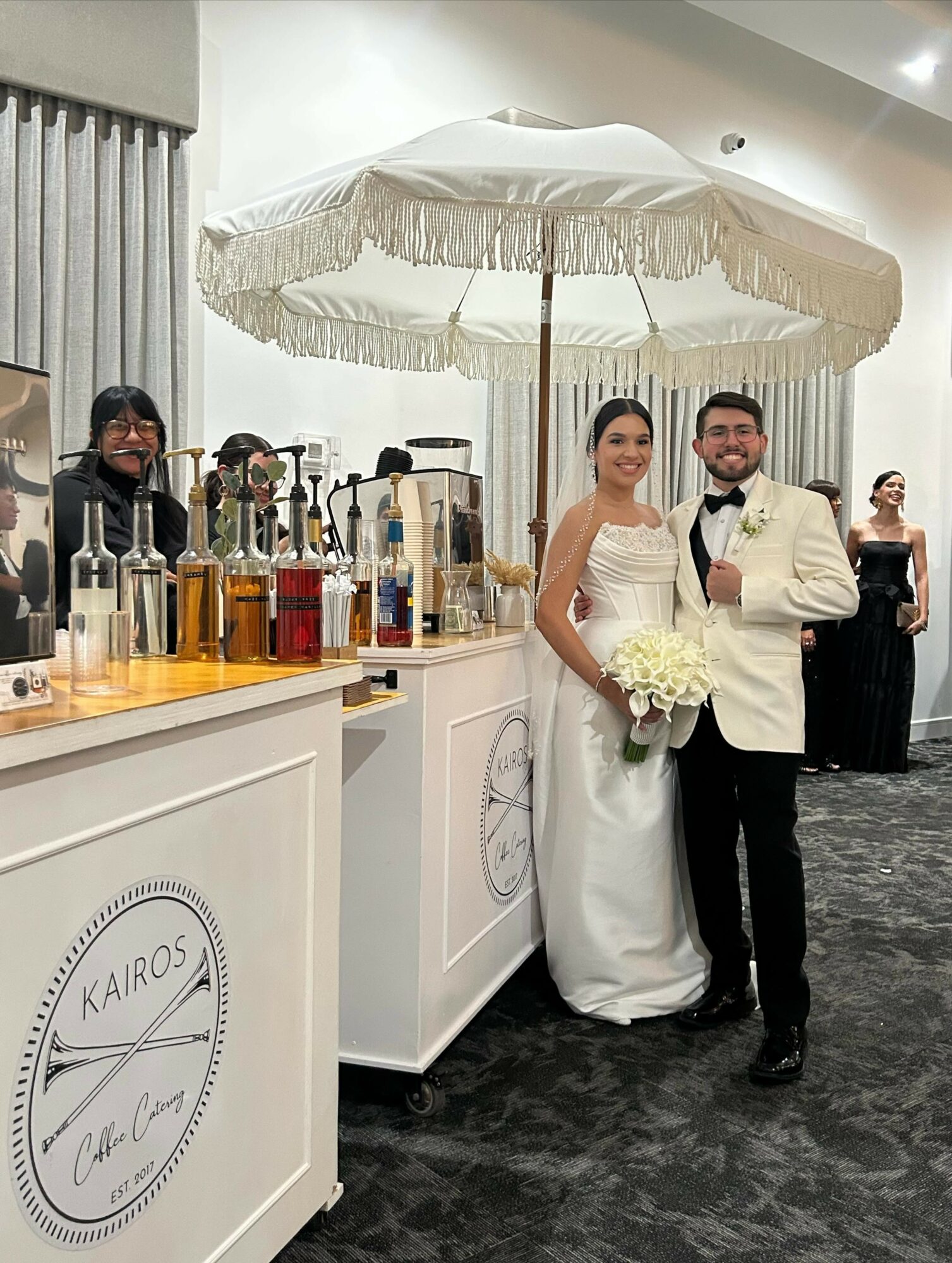 Bride and groom in wedding attire under a large white umbrella, standing beside a bar with bottles, in a decorated indoor venue.