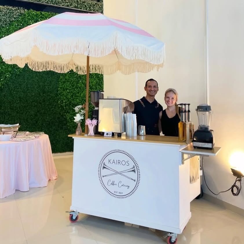 Two people stand behind a white mobile bar with a logo, under a pink and white umbrella, at an outdoor event.