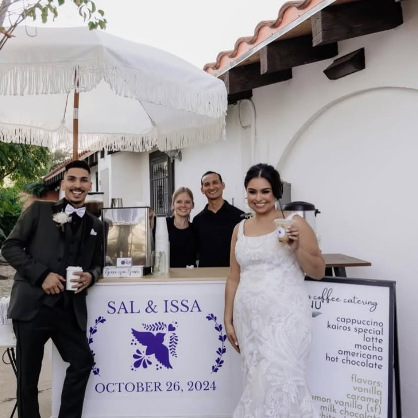 Four people at a wedding reception outdoors, with a white table, umbrella, and white building in background.