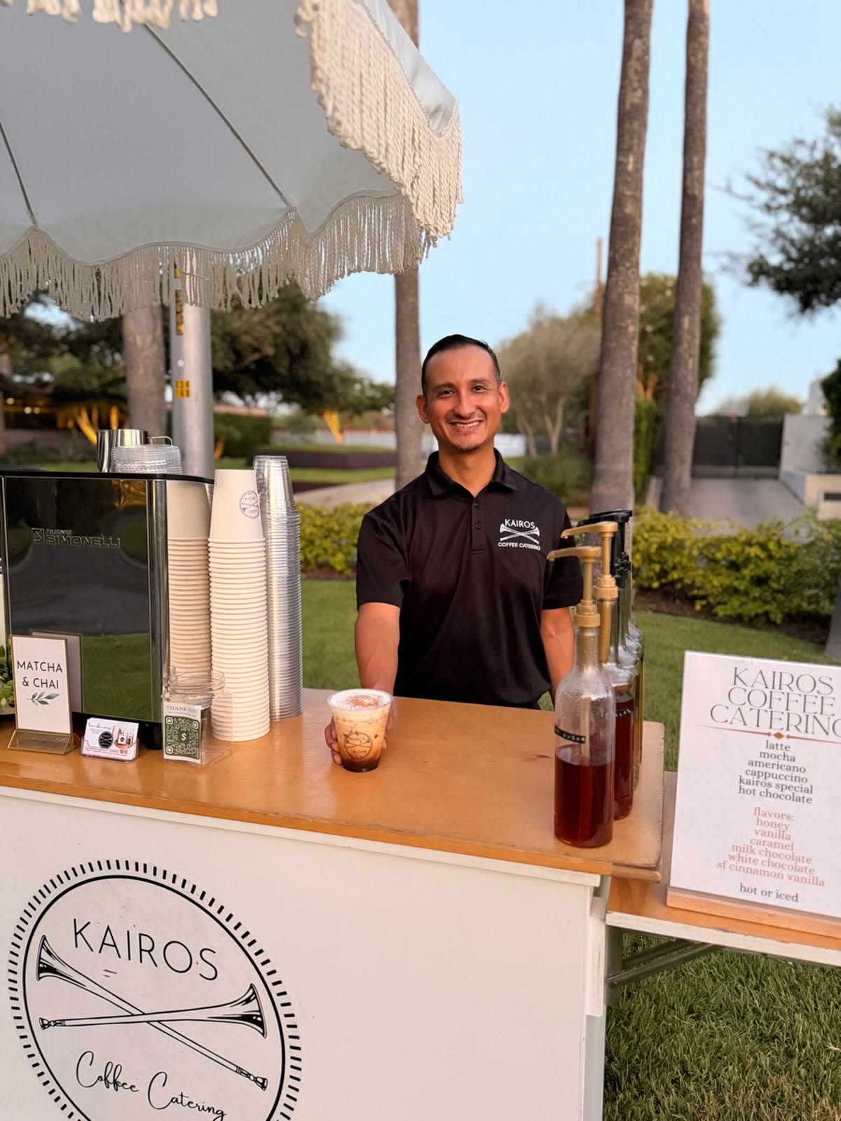 Smiling man in black shirt behind a coffee stand outdoors with trees and blue sky in background.