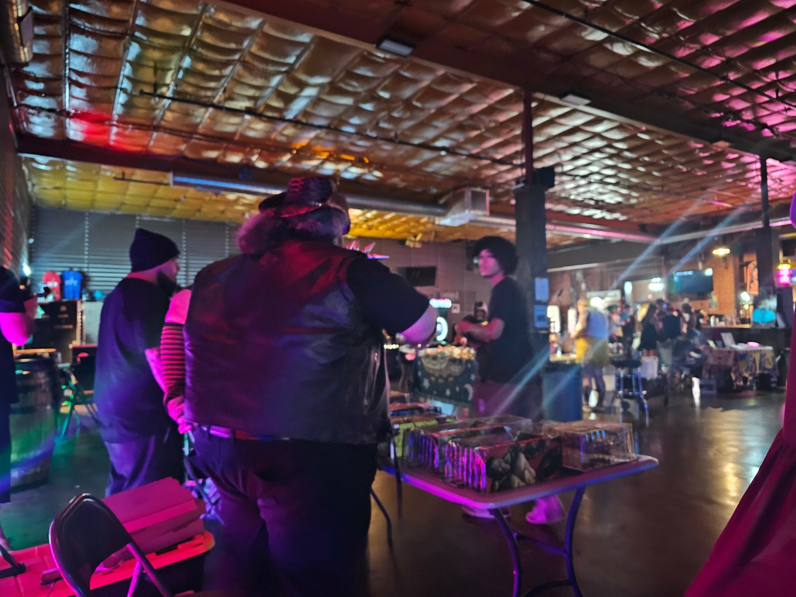 People gather around a table with items in a dimly lit indoor space with colorful lighting.