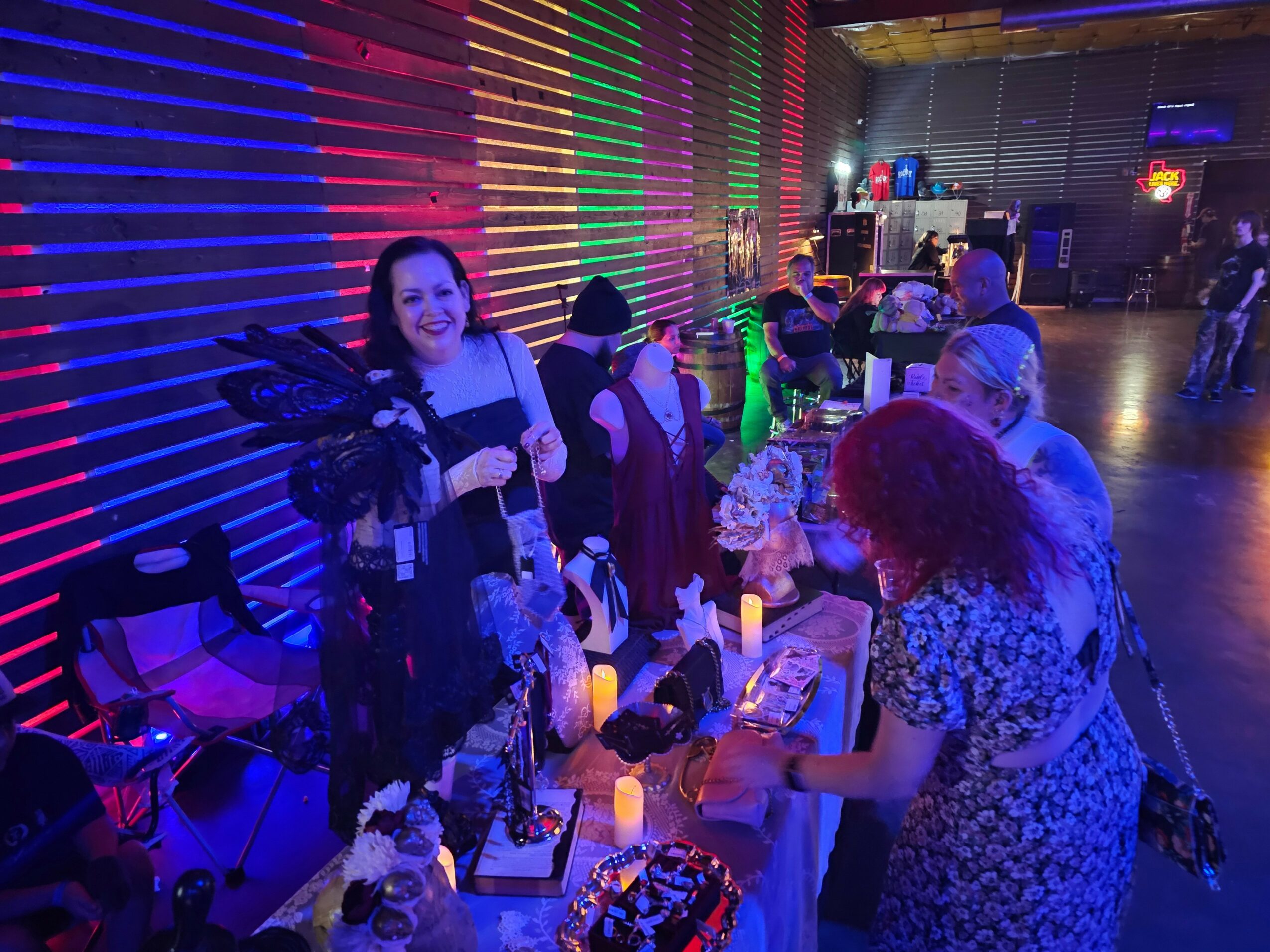 Group of people at a colorful indoor event, some browsing tables with items, illuminated by vibrant neon lights.