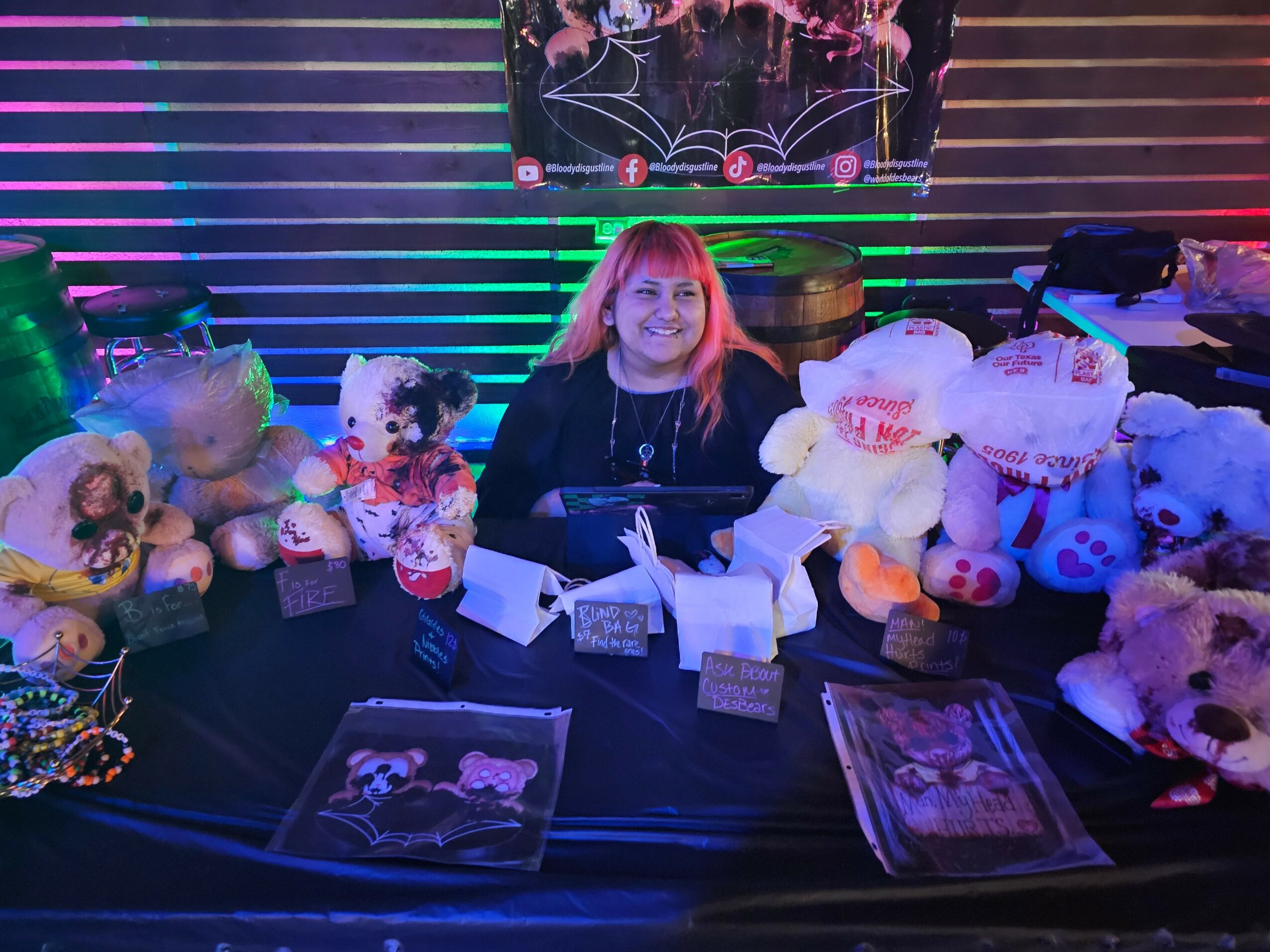 Woman sitting at a table with stuffed animals and books, colorful striped background, smiling.