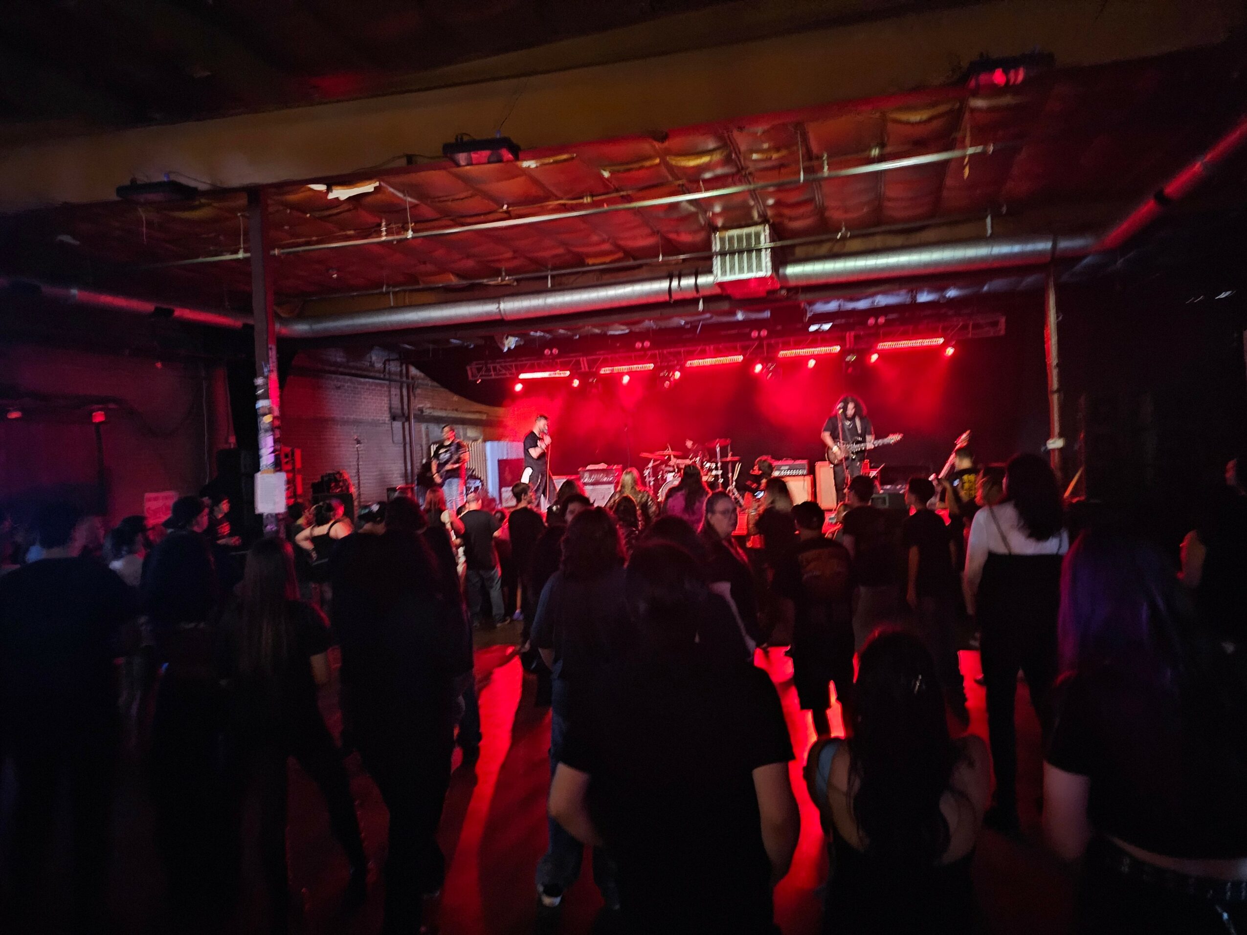 Crowd watching a live band perform on stage with red lighting in a dark indoor venue.