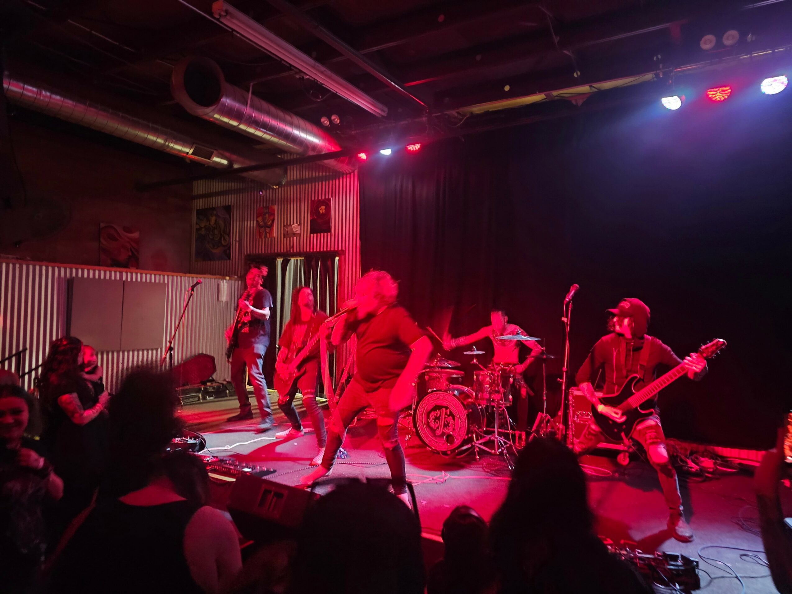 Band performs on stage with red and blue lighting, audience watching in a dark venue.