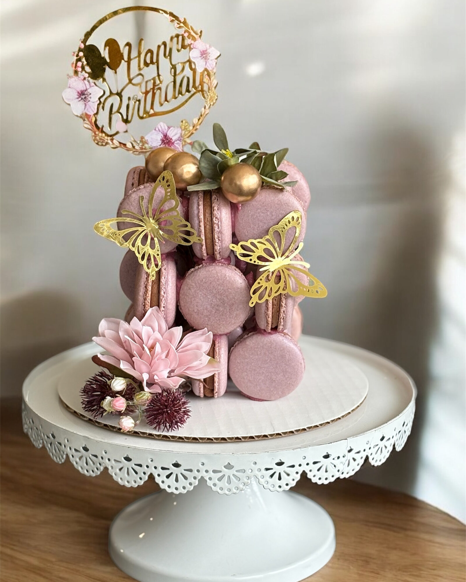 Pink macarons decorated with gold butterflies, flowers, and a 'Happy Birthday' topper on a white cake stand.