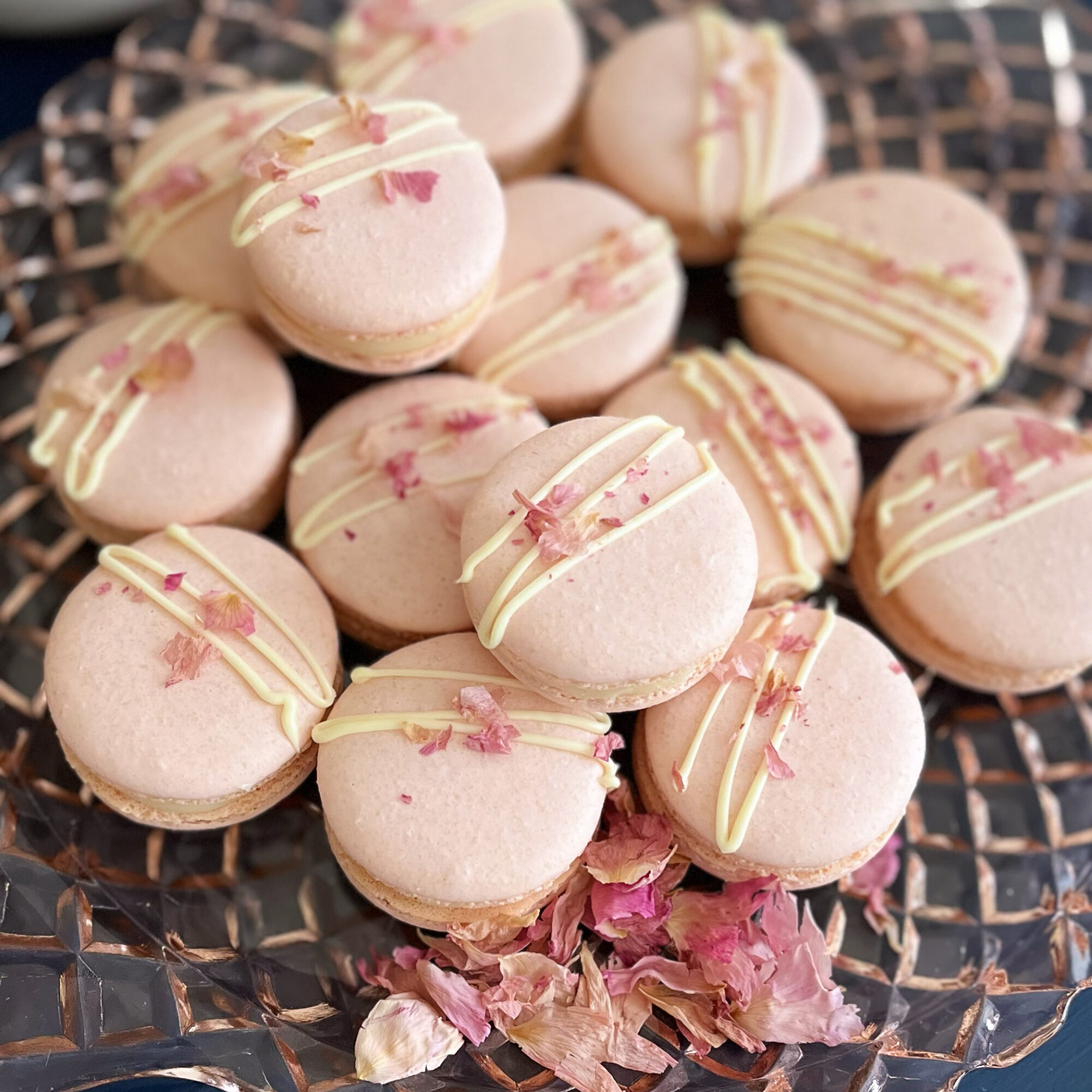 Pink macarons with white and pink drizzle, garnished with pink flower petals, on a wire rack, some petals scattered below.