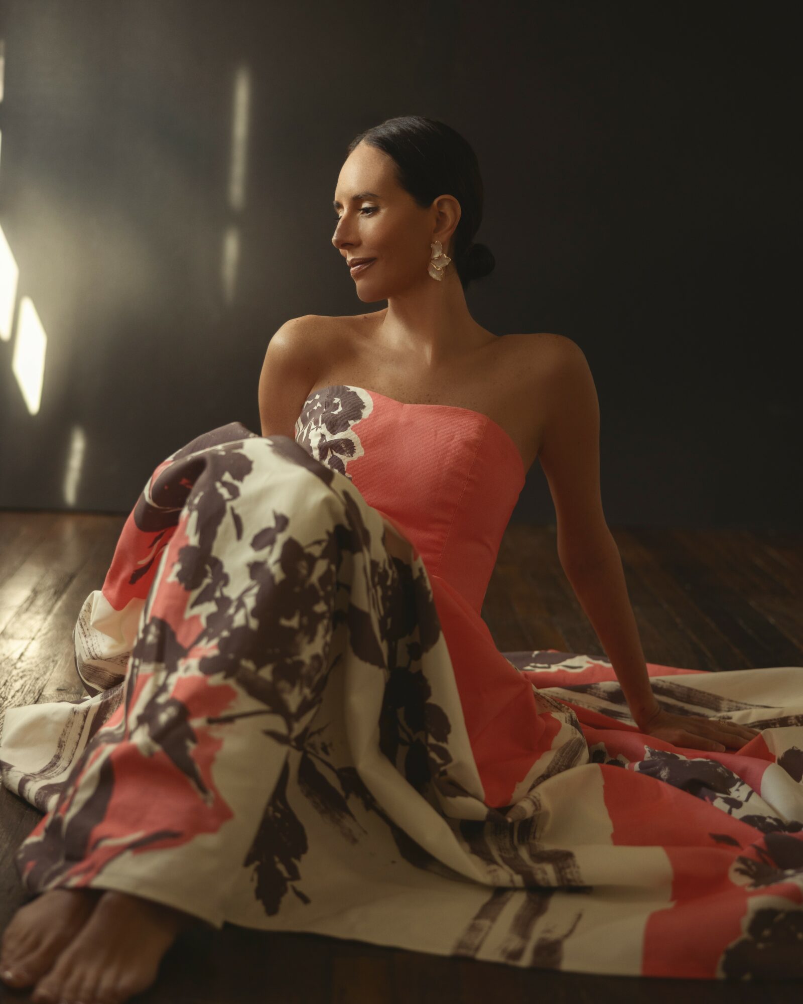 Woman in a strapless pink dress sitting on the floor with a floral blanket, looking to the side, dim lighting.